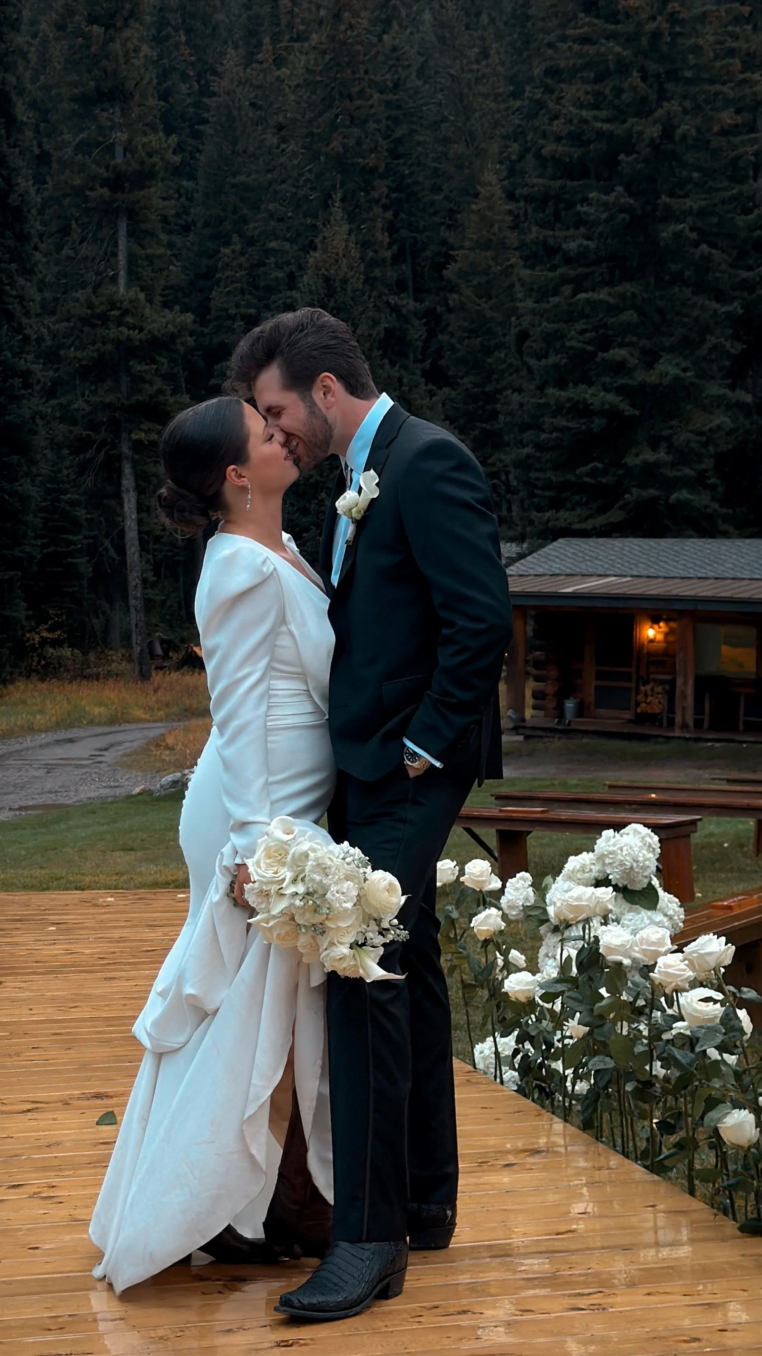 A bride and groom sharing a kiss on a wooden outdoor platform decorated with white flowers in front of a forested background during a wedding ceremony.