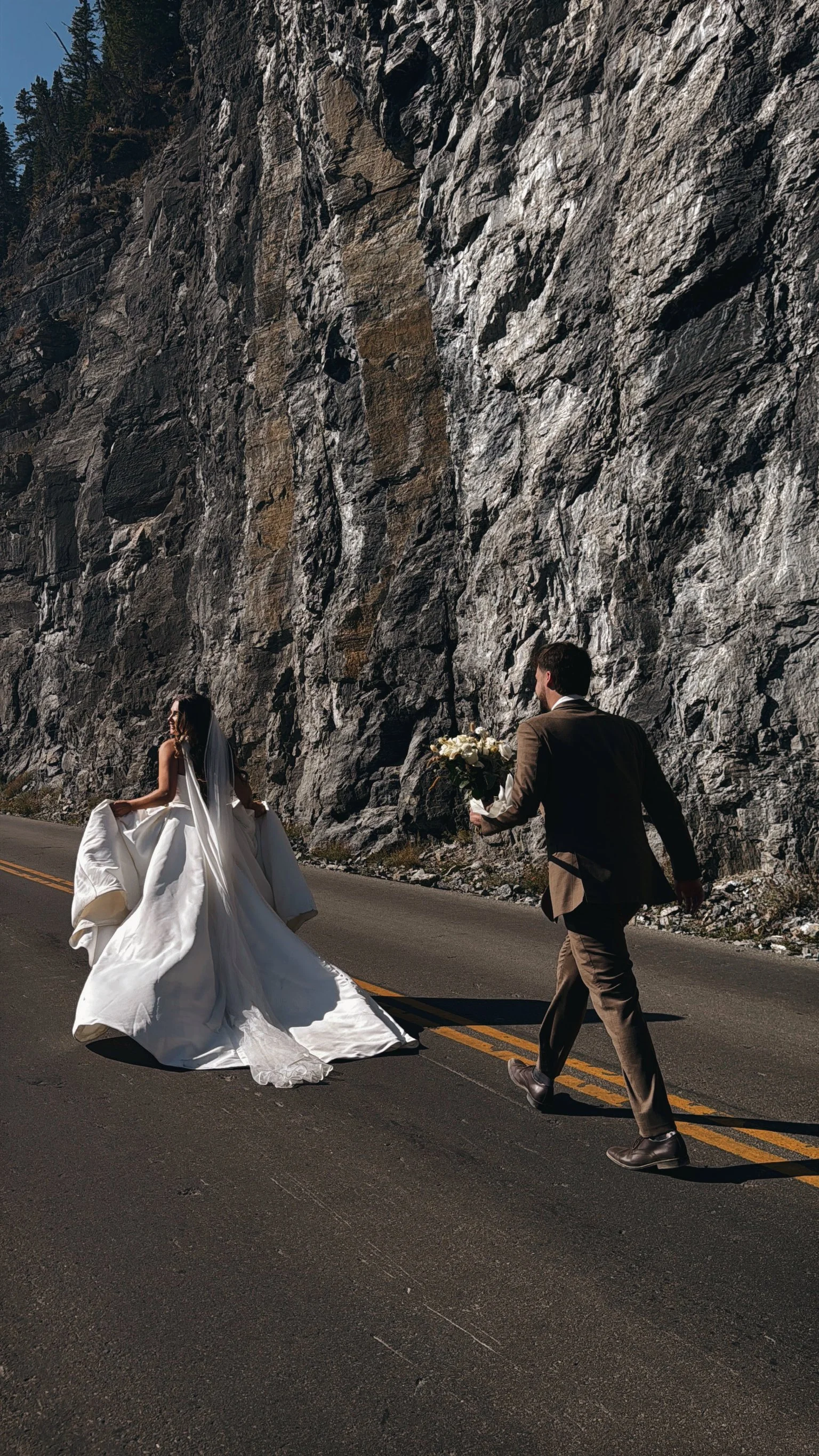 A bride in a white wedding dress and veil is walking on a road with a groom in a suit approaching with a bouquet of flowers, next to a large rocky mountain at what appears to be sunset or sunrise.