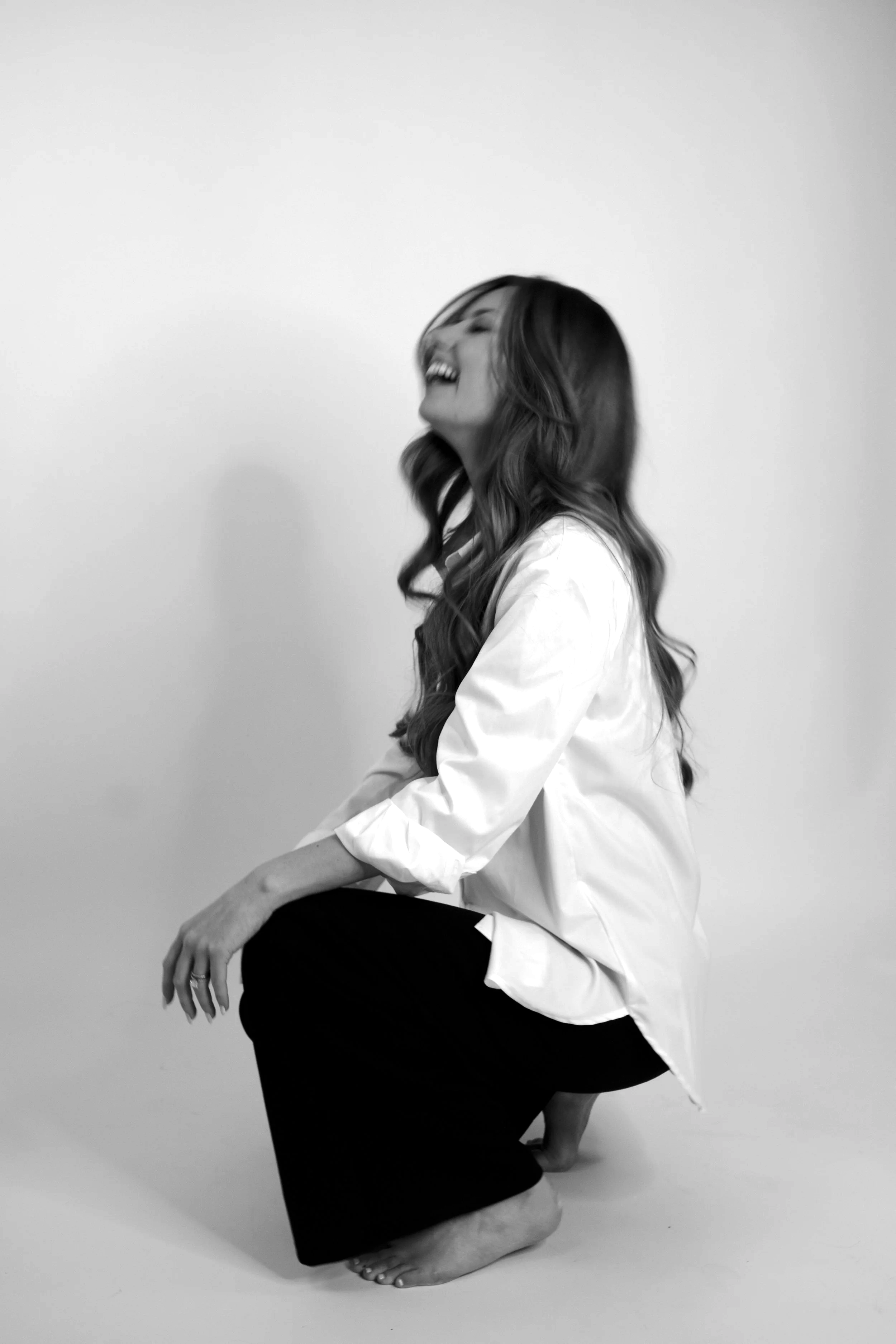 A woman with long wavy hair is sitting on the floor against a plain backdrop. She is smiling and appears to be laughing, wearing a white shirt and black pants.