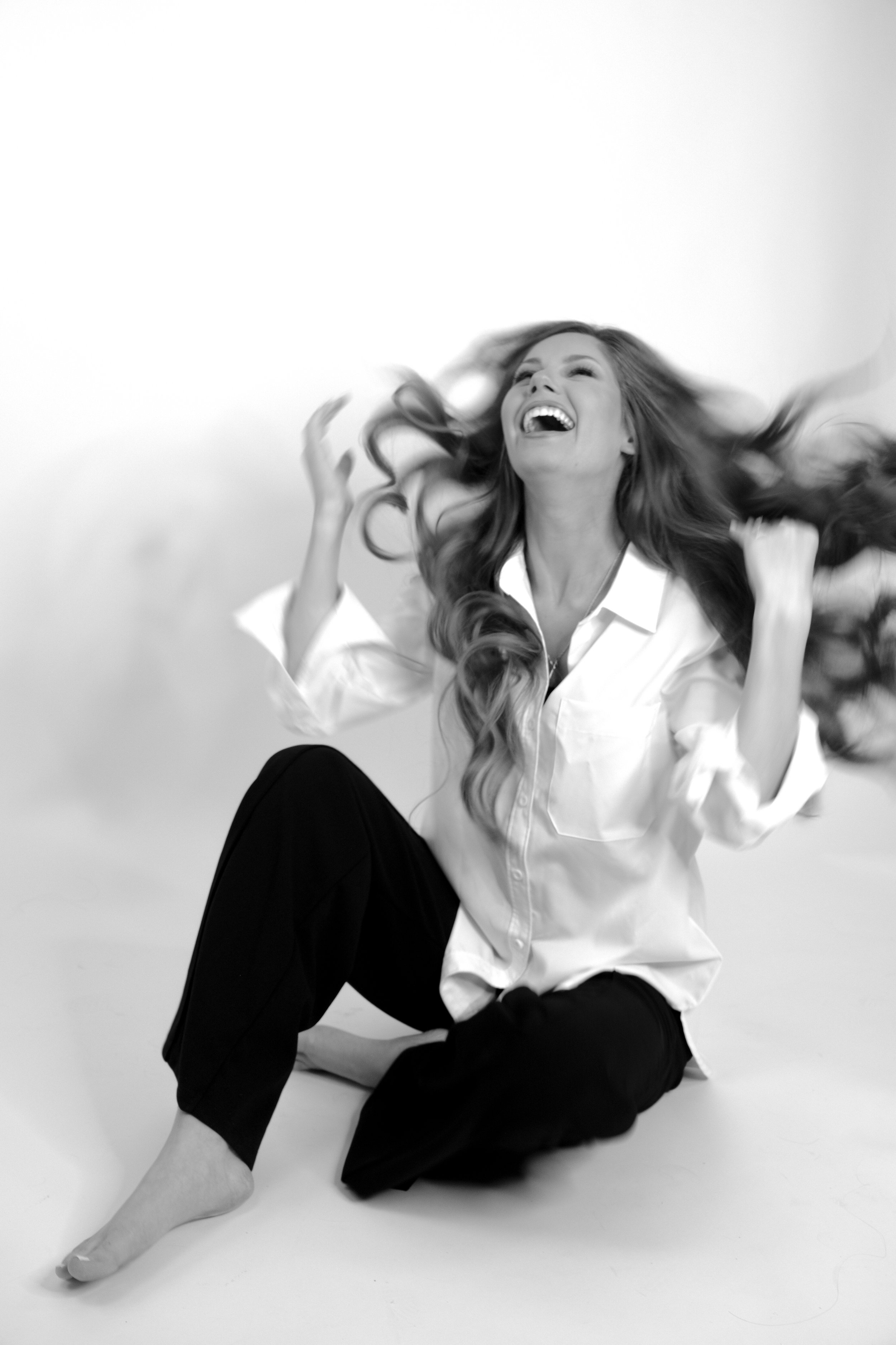 A woman with long hair sitting on the floor, laughing joyfully, wearing a white shirt and black pants, in a studio setting with a plain background.