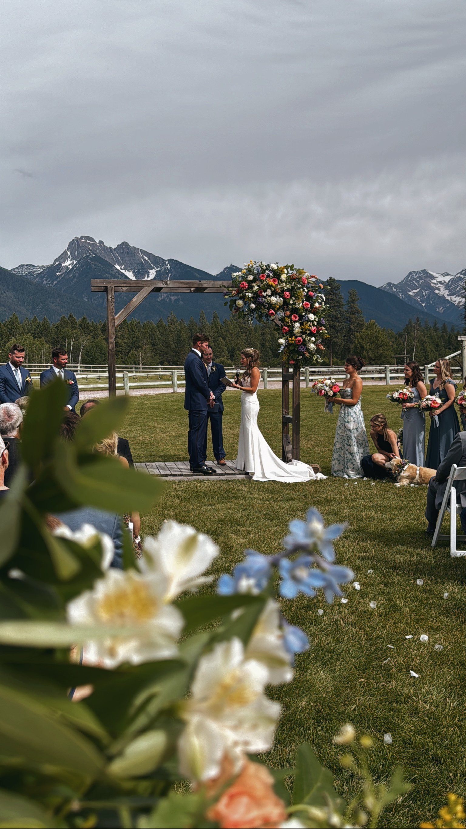 A wedding ceremony taking place outdoors with mountains in the background. The bride and groom are facing each other under a floral arch, with bridesmaids and groomsmen present. The scene is surrounded by greenery and colorful flowers.