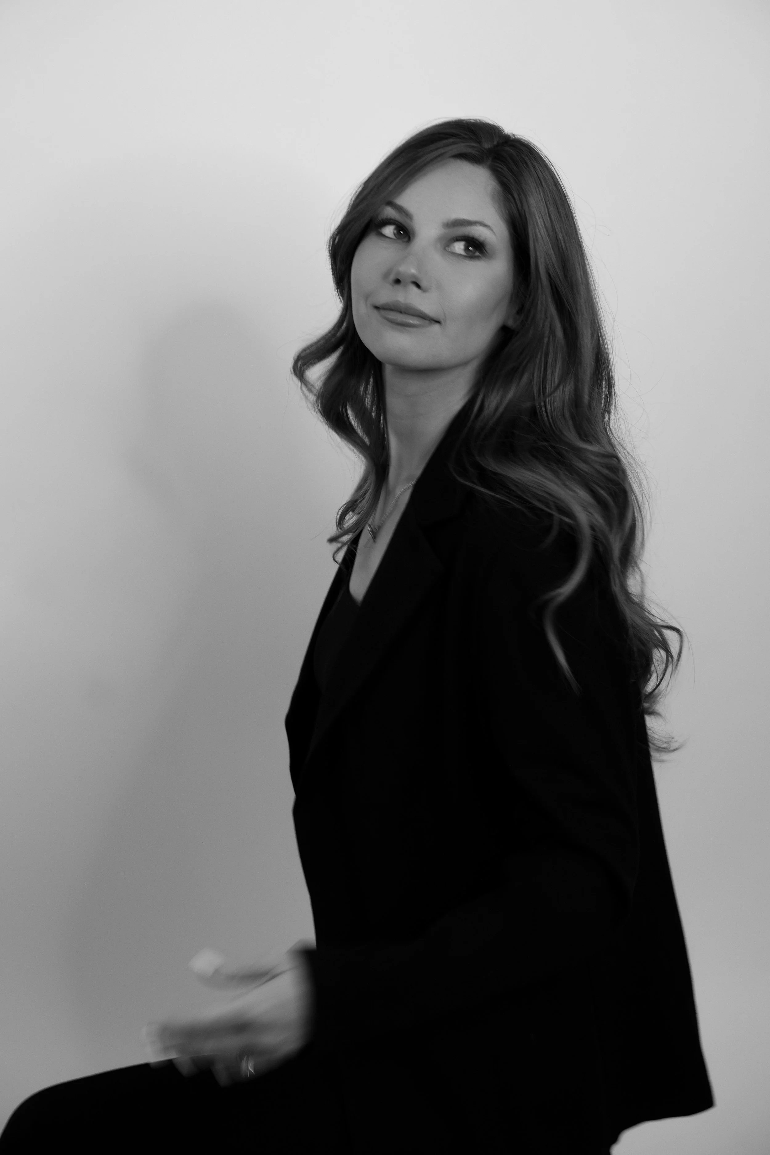 Black and white portrait of a woman with long wavy hair, looking to her left, wearing a blazer, against a plain background.