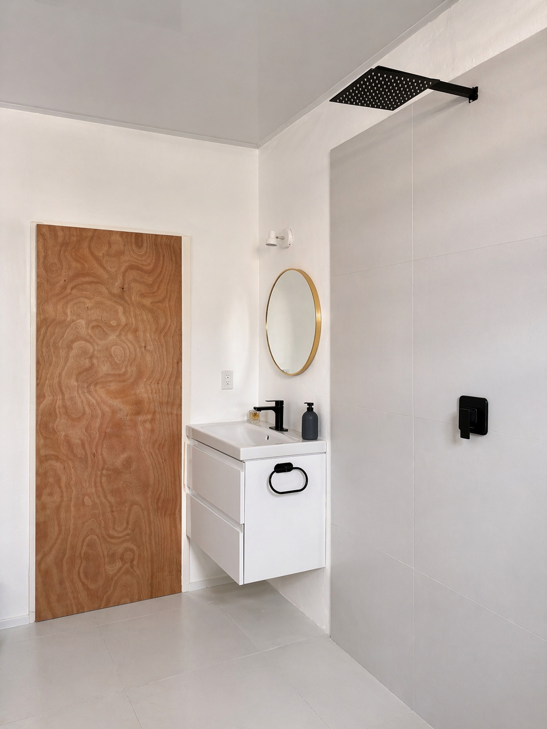 Modern bathroom with white walls, a wooden door, a white sink cabinet with a black faucet, a round mirror with a gold frame, a black soap dispenser, and a black wall-mounted showerhead.