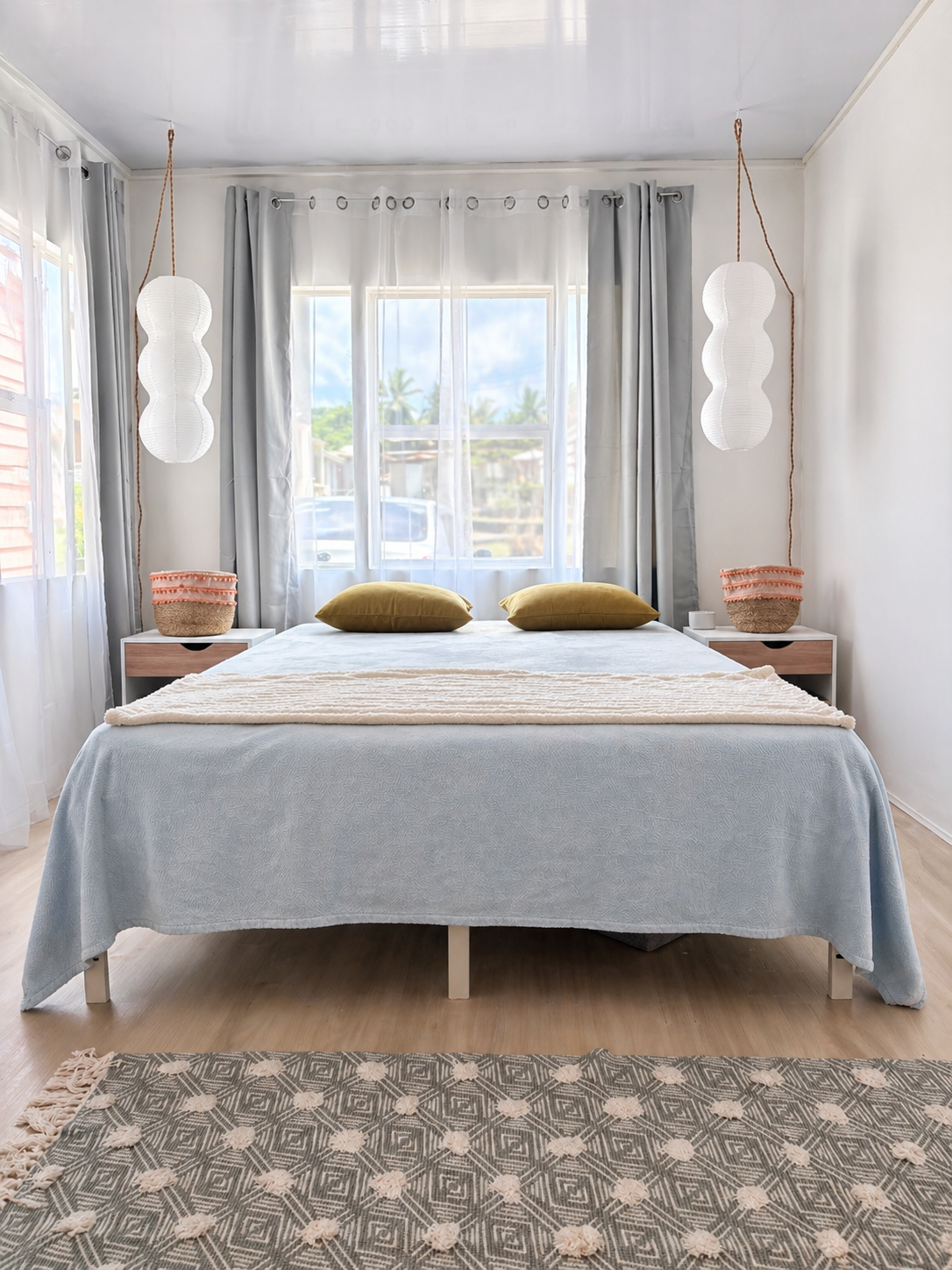 A bedroom with a bed, two pillows, two nightstands, hanging pendant lights, and a window with curtains, overlooking a street with cars and palm trees.