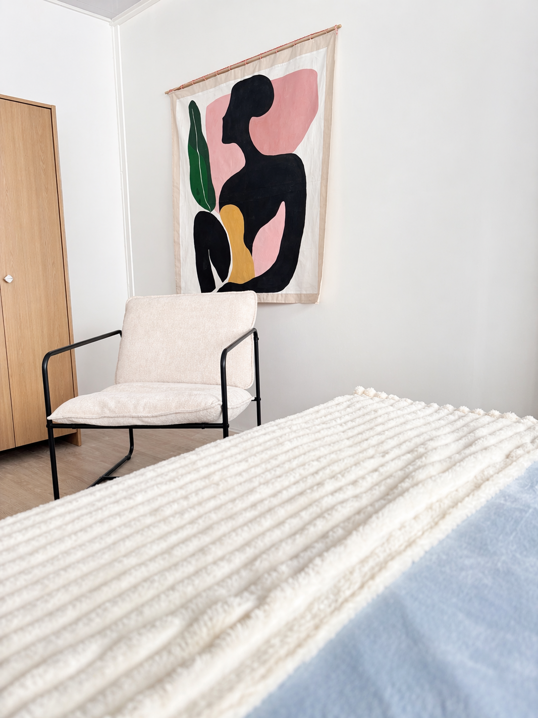 A minimalist bedroom featuring a white textured bedspread, a beige cushioned chair with black metal arms, a wooden wardrobe, and a modern abstract wall art depicting a silhouette of a woman with colorful geometric shapes and a green leaf.