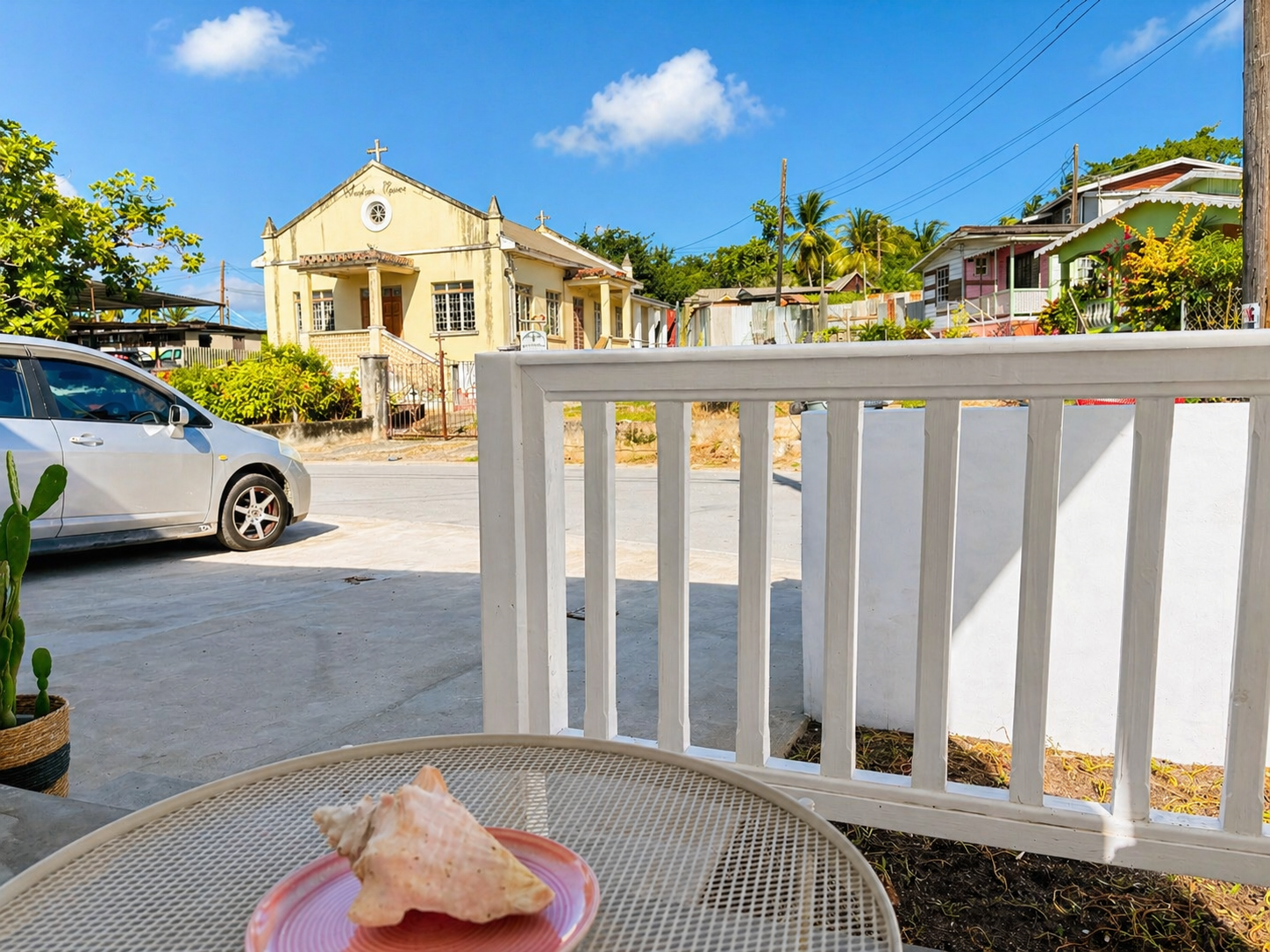 View of a street with a church, colorful houses, a parked car, and a potted cactus; a table with a large seashell in the foreground.