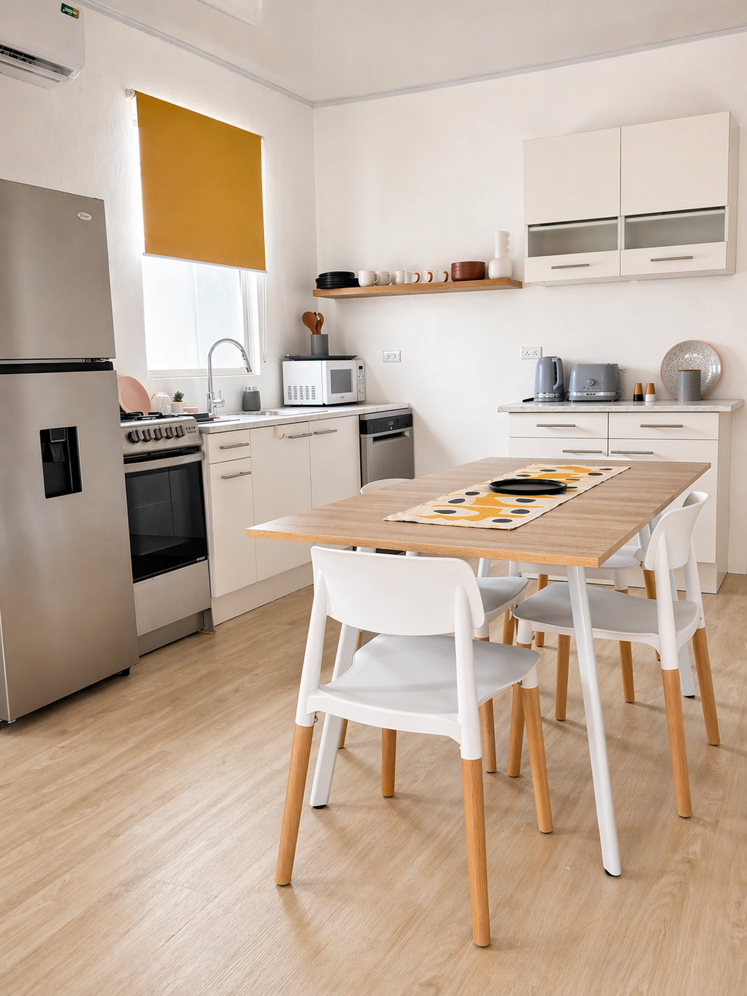 Modern kitchen with white cabinets, wooden dining table with white chairs, stainless steel refrigerator, microwave, toaster, kettle, dishware, and a window with a yellow blind.