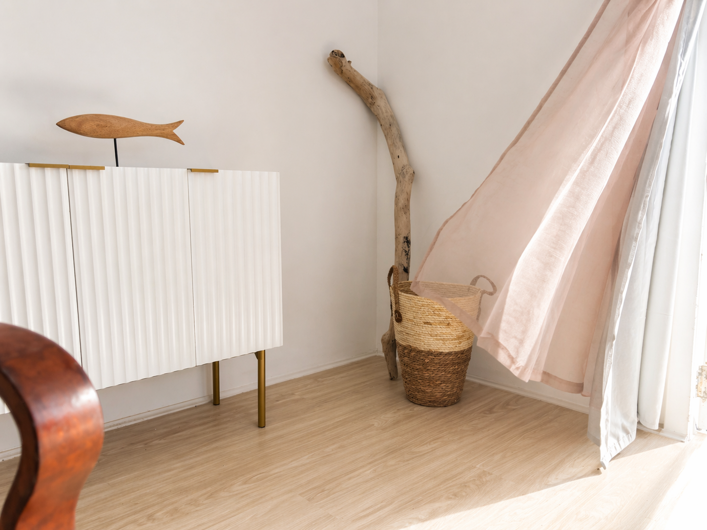 Corner of a room with a wooden fish sculpture on a white cabinet, a piece of driftwood leaning against the wall, woven baskets on the floor, and light pink curtains partially covering a window.