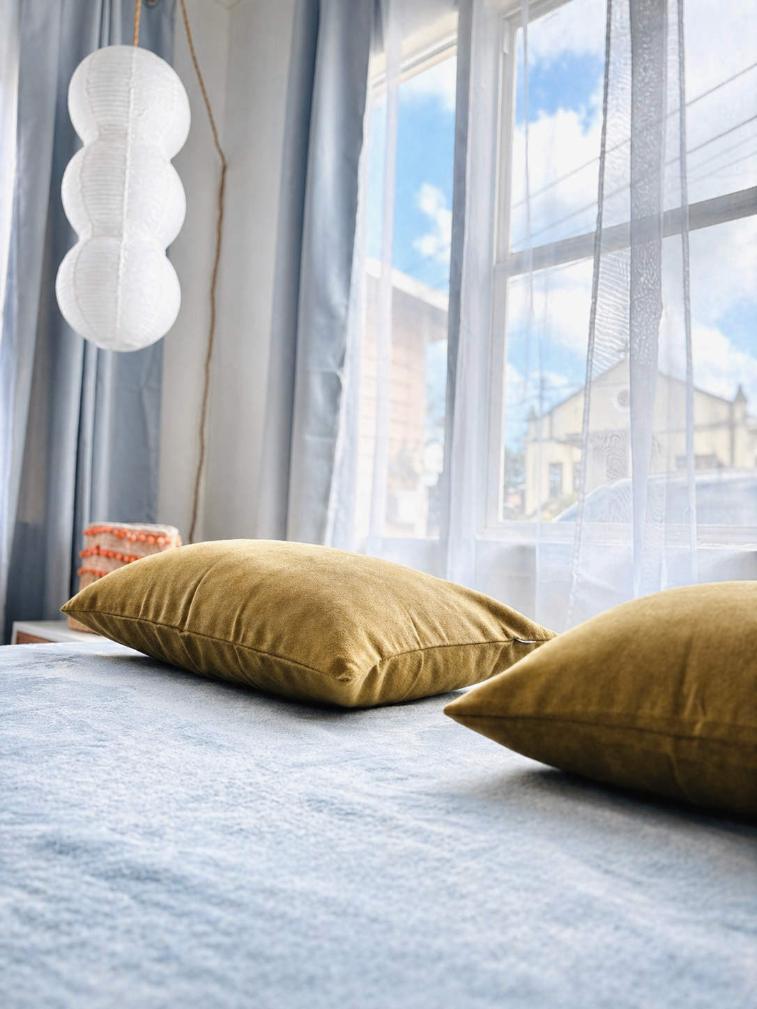 Sunlit bedroom with two gold pillows on a bed, sheer curtains, a paper lantern, and a window showing a blue sky and houses outside.