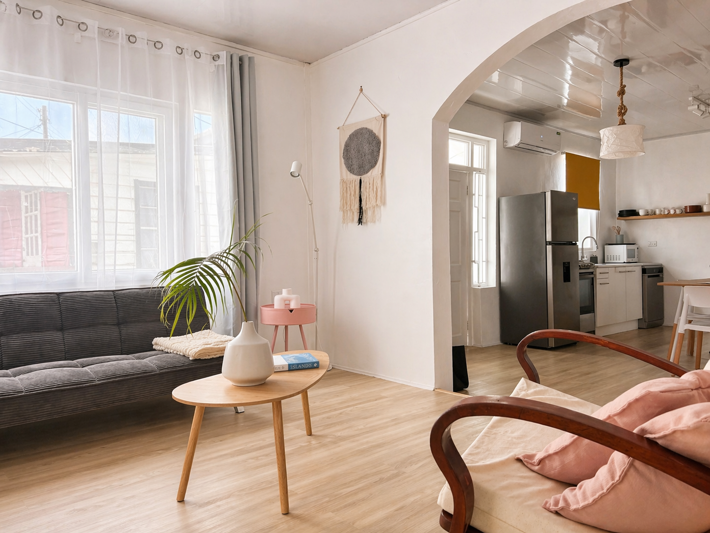 Living room with large window, dark couch, round wooden coffee table with vase and book, pink side table, light-colored wood floor, white walls with wall hanging, adjacent open kitchen with refrigerator, microwave, and dining table.