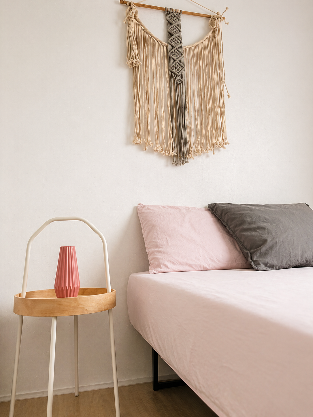 A bedroom corner with a bed that has pink and gray pillows, a wooden side table with a pink vase, and a decorative macramé wall hanging above the bed.