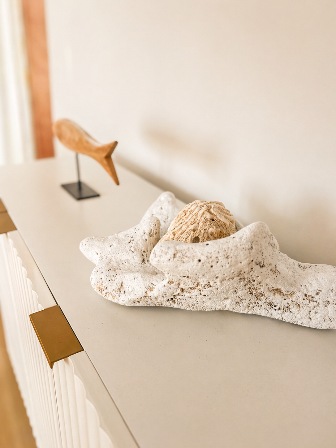 Decorative display with a large white coral shell holding a textured beige seashell, with a wooden fish sculpture on a stand in the background.