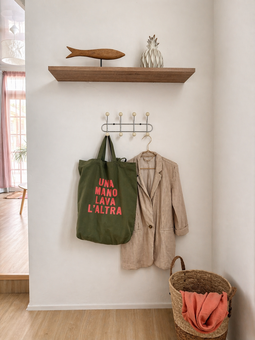A wall with a wooden shelf and a decorative wooden fish and ceramic pineapple on top, a hook with a green tote bag and striped blazer hanging, a woven basket with an orange cloth underneath.