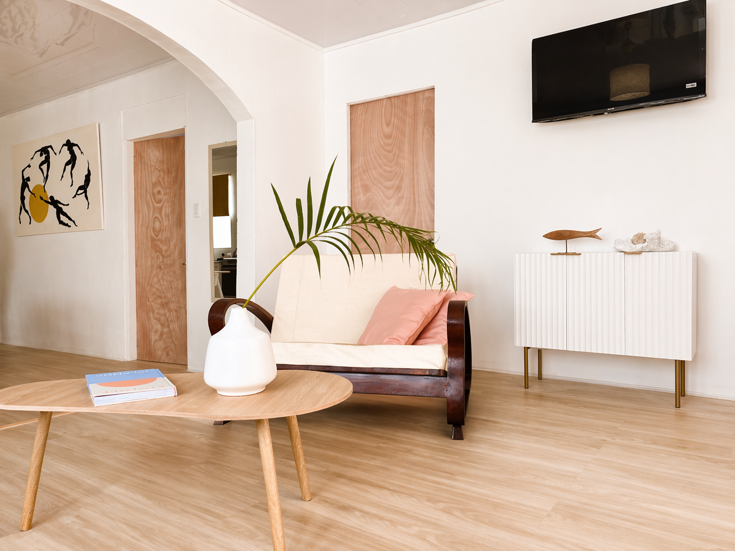 A living room with a white wall, flat-screen TV, modern white cabinet with gold legs, a wooden fish sculpture, artwork of dancing figures, a beige sofa with pink pillows, a light wooden coffee table with a white vase and green plant, hardwood flooring, and an arched ceiling.
