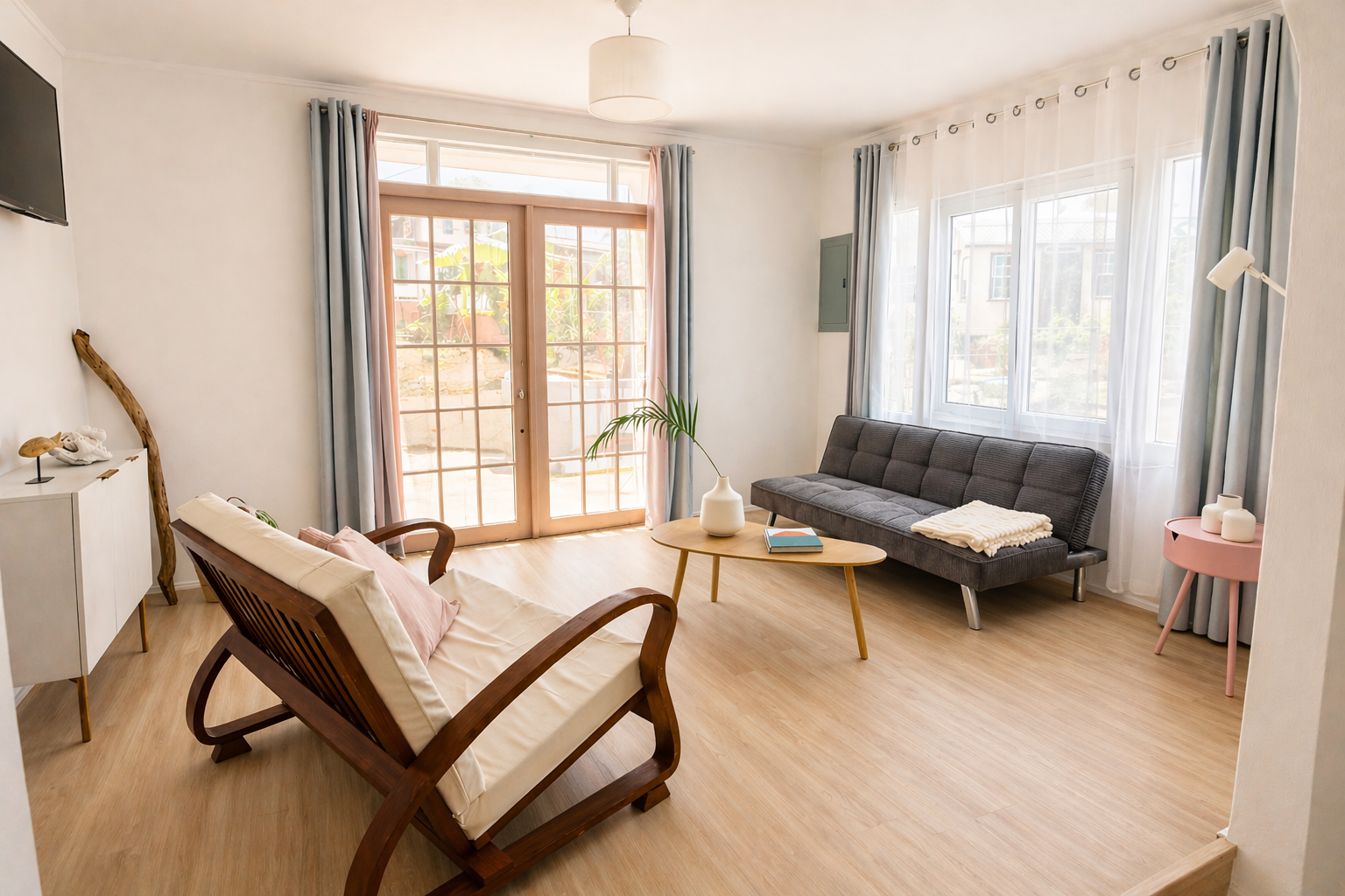 Living room with wooden floor, beige walls, large windows with light blue curtains, sliding glass door, dark gray sofa, wooden coffee table with a white vase and books, wooden armchair with white cushion and pink pillow, white sideboard with decorati