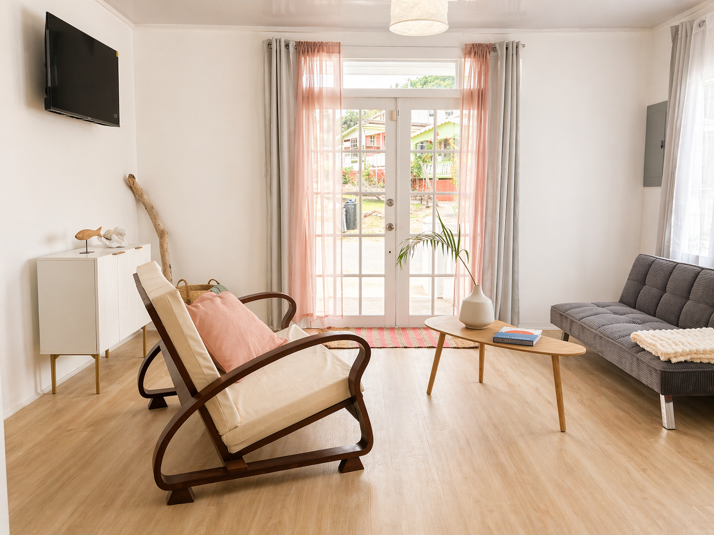 Bright living room with a sliding glass door covered by pink and white curtains, wooden floor, modern gray sofa, wooden coffee table with a white vase and book, cushioned armchair with pink pillow, TV on the wall, and natural decorations.