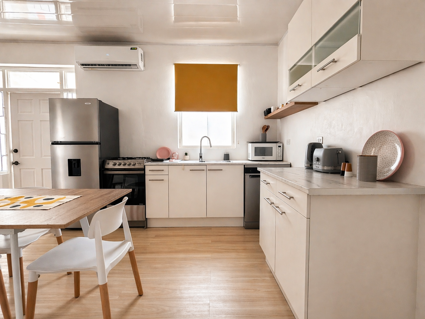 Modern kitchen with white cabinets, stainless steel refrigerator, oven, microwave, and small appliances on countertop. Wooden dining table with white chairs. Window with yellow shade, wooden floor, white walls.