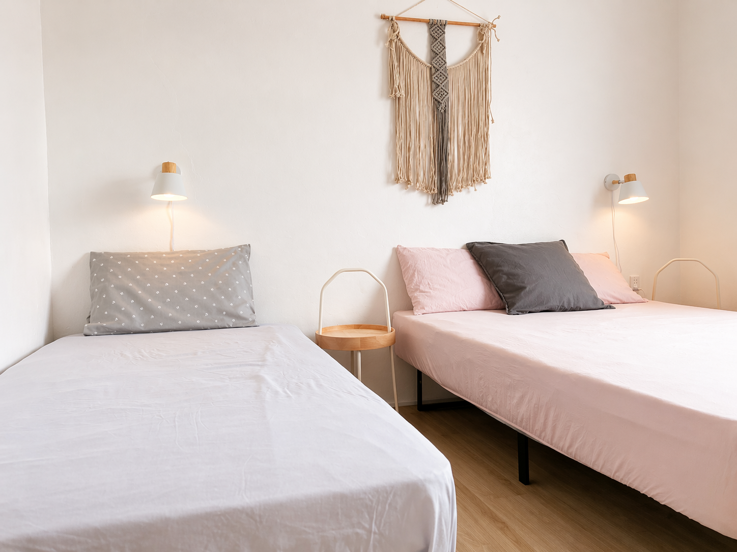 Two beds in a minimalist bedroom with wall-mounted reading lamps, a small wooden side table between them, and a decorative hanging wall art above.