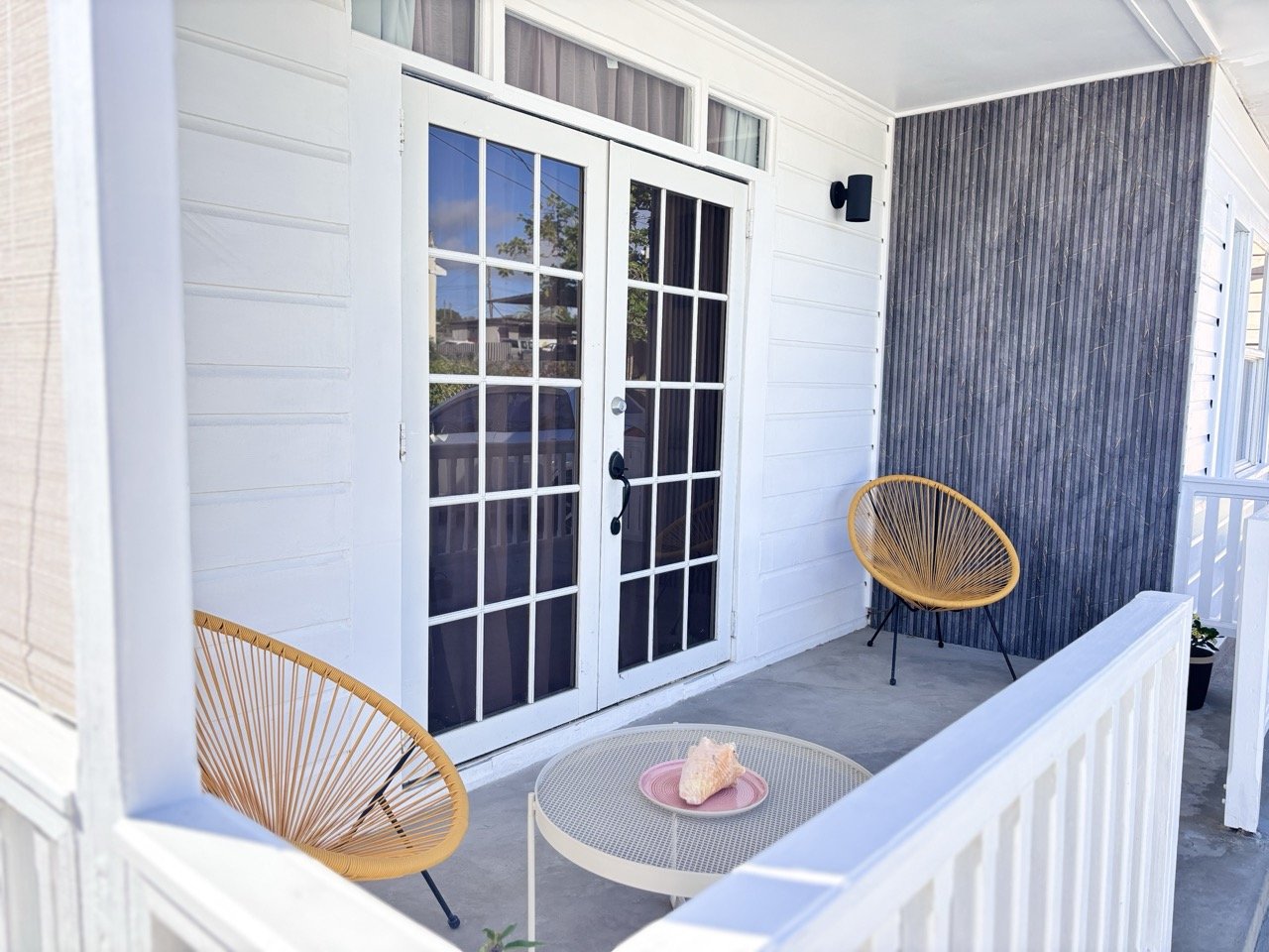 A porch with white siding, glass double doors, two yellow round chairs with black legs, a small white round table with a pink plate and a light-colored shell, and a small potted plant on the railing.