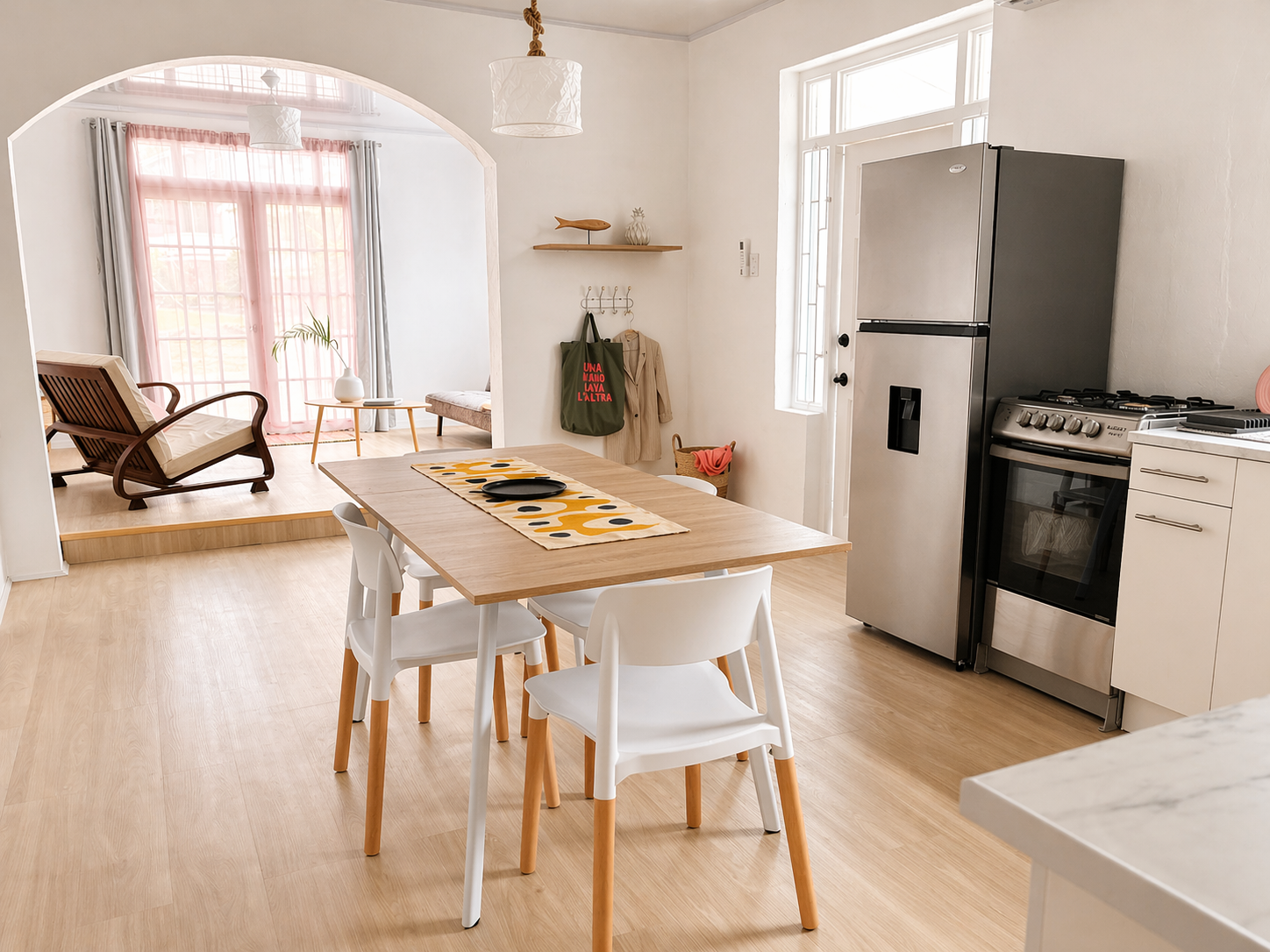 Bright kitchen and dining area with wooden table and six white chairs, stainless steel refrigerator, oven, and gas stove, with a cozy sitting area in the background featuring armchair and sofa, decorated with pink curtains and natural light.