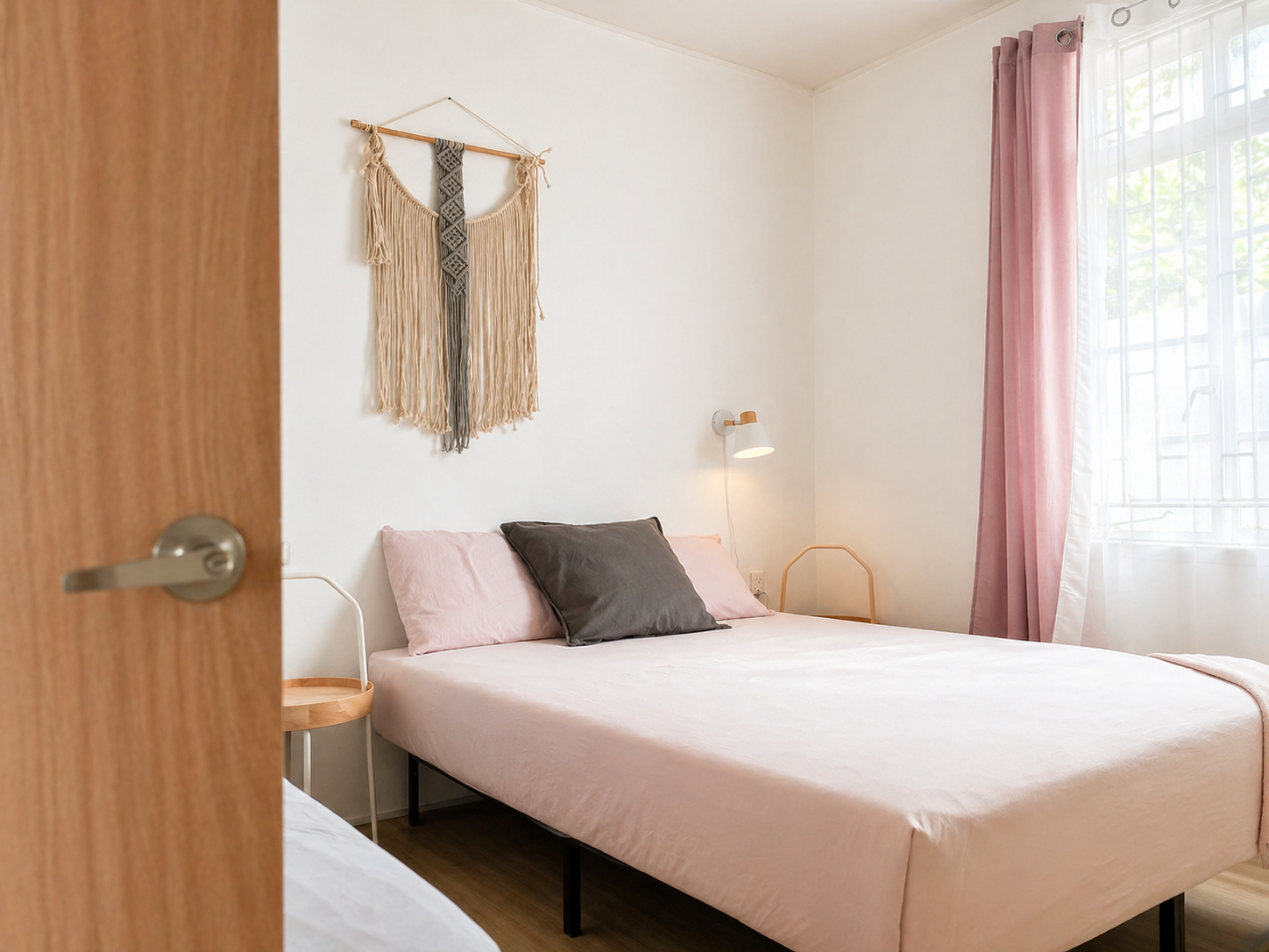 A bedroom viewed partially through a doorway, with a bed made up with pink bedding and cushions, a wall-mounted lamp, pink curtains, a window with bars, and a bohemian wall hanging.