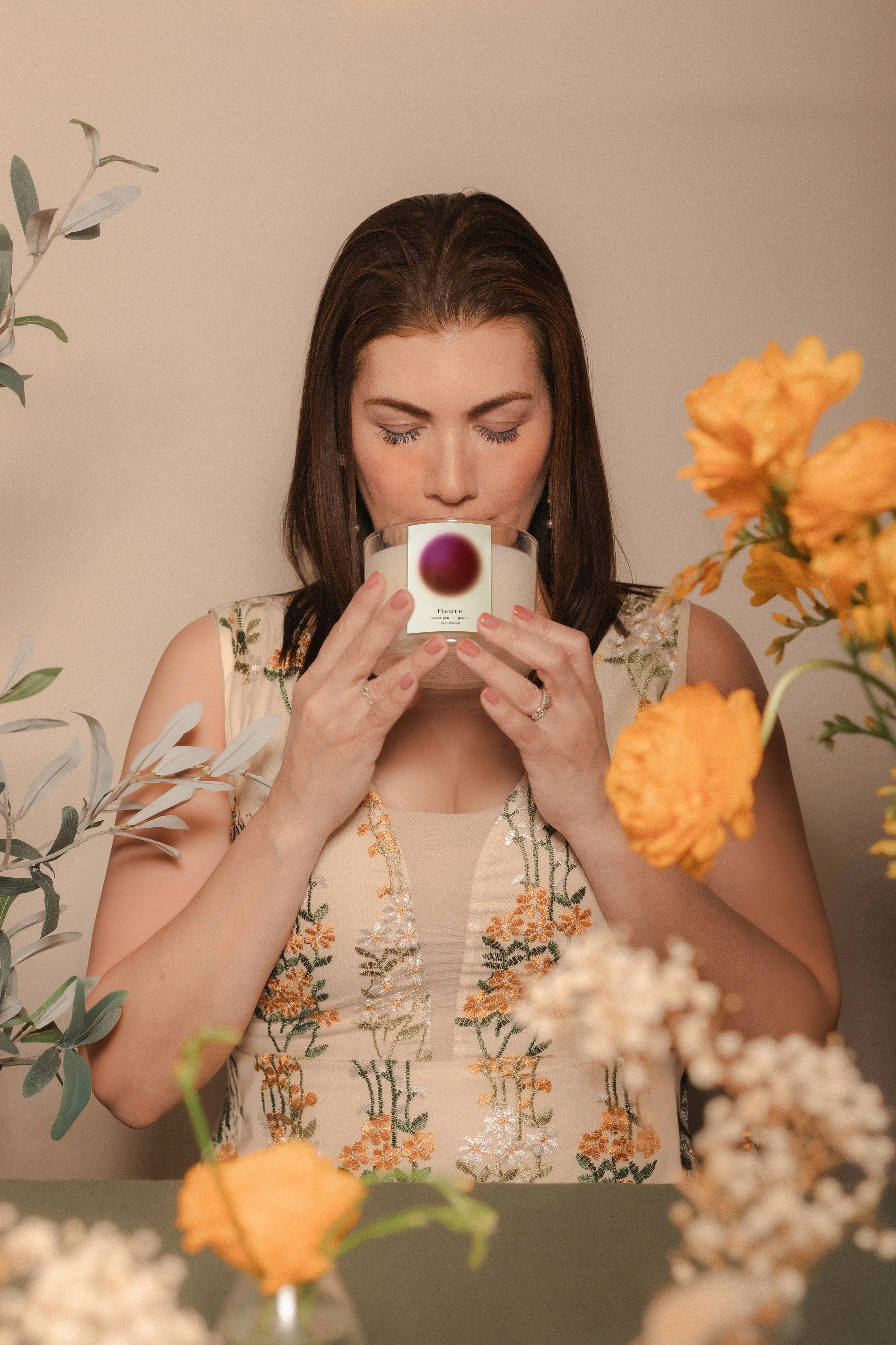 Our founder, a woman with dark brown hair and light skin holding a candle with a purple dot on it, surrounded by flowers and greenery, wearing a floral embroidered dress.