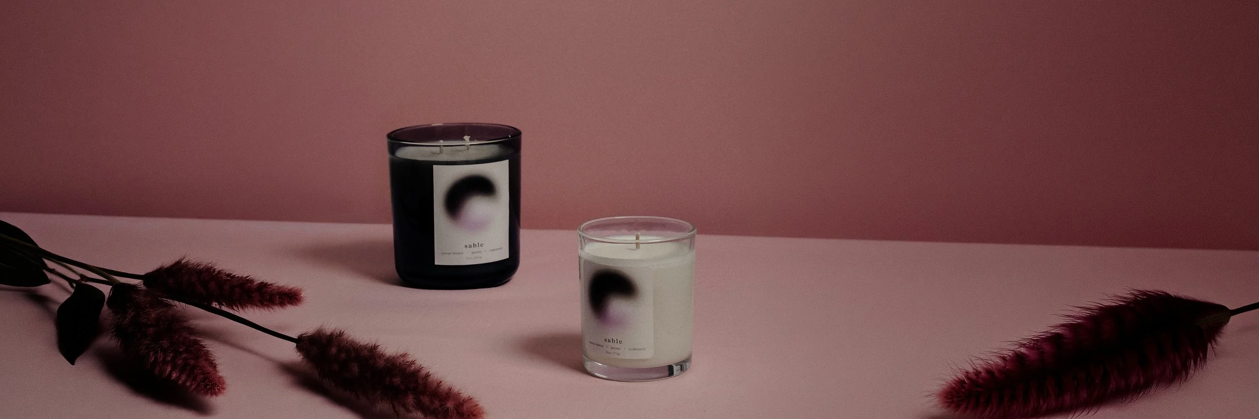 Two candles in black and clear glass holders with labels featuring a blurred circle design, placed on a pink surface with pink fluffy plants on either side, against a pink background.