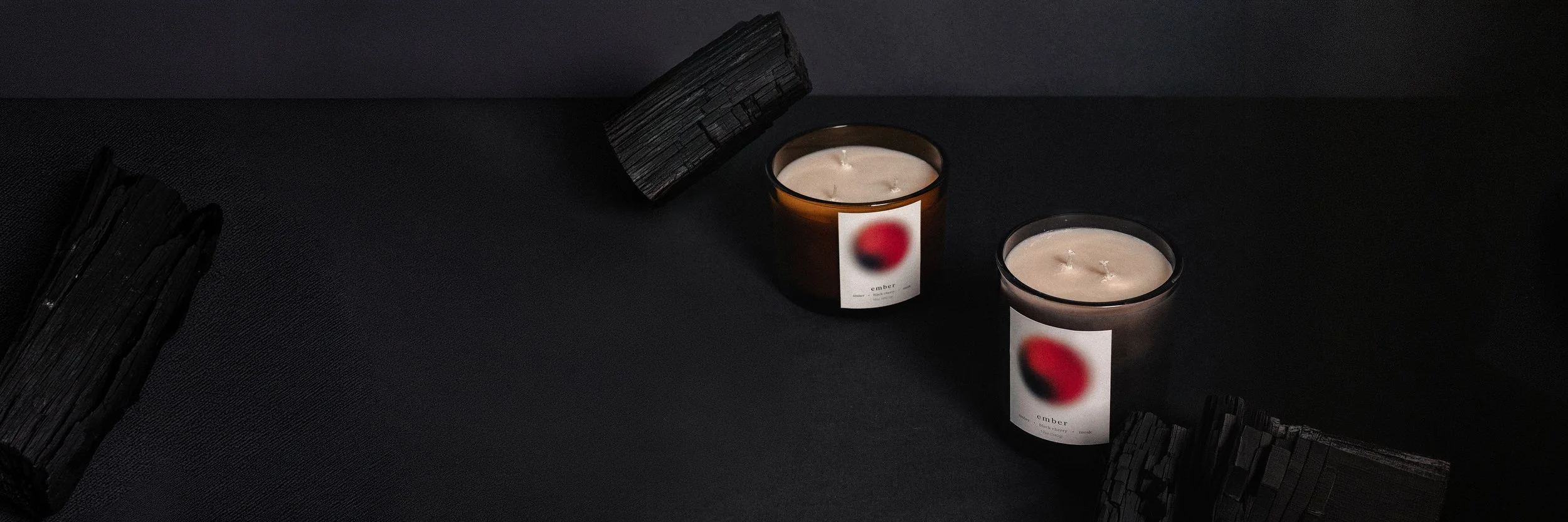 Two scented candles in dark brown glass containers with white labels featuring red and black blurred circular designs, surrounded by black burned wood pieces, on a black textured surface.