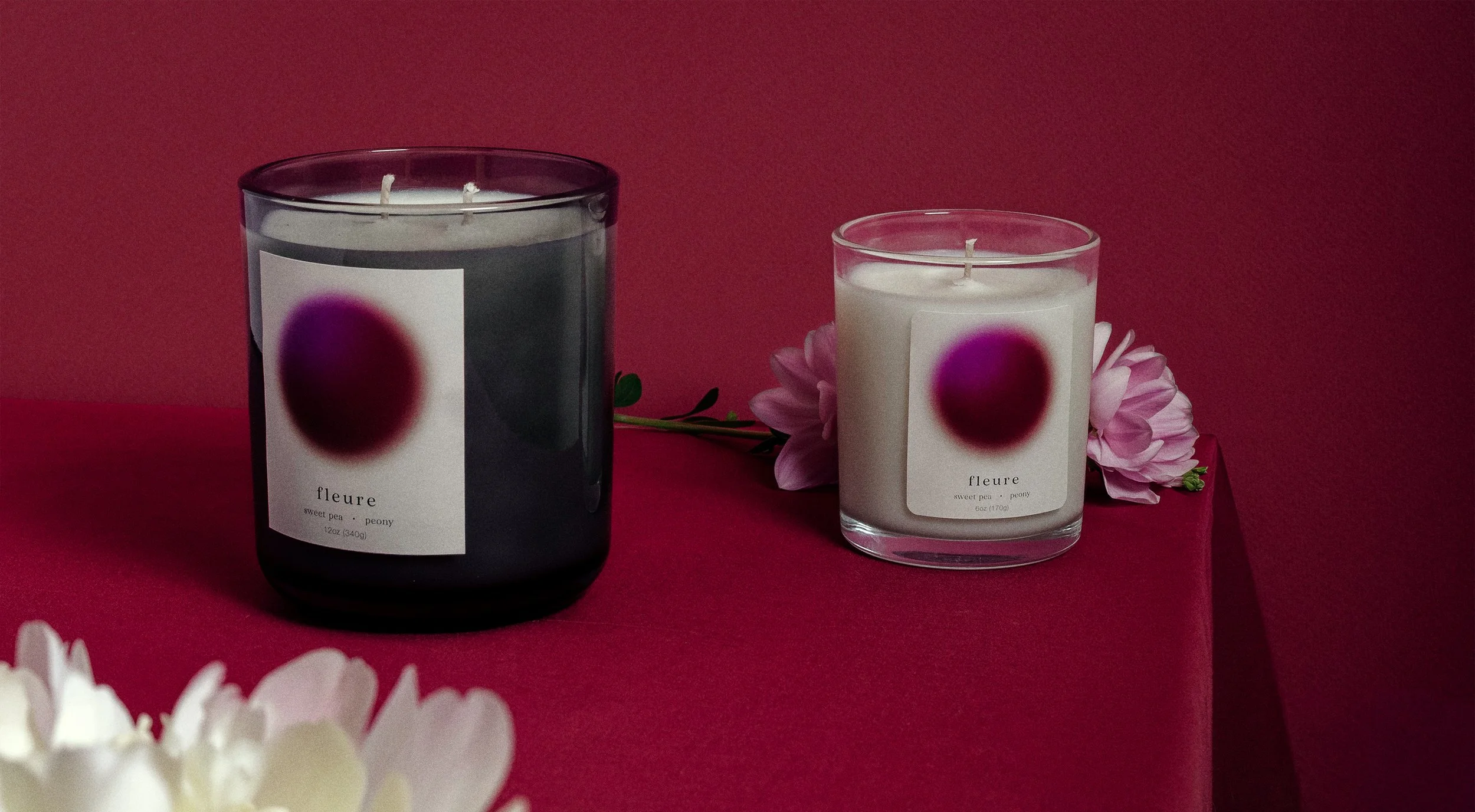 Two candles in glass jars, one dark gray and one white, with labels reading 'fleure, sweet pea & peony'. The candles are placed on a magenta surface, with pink flower petals and white flowers nearby, against a monochromatic background.
