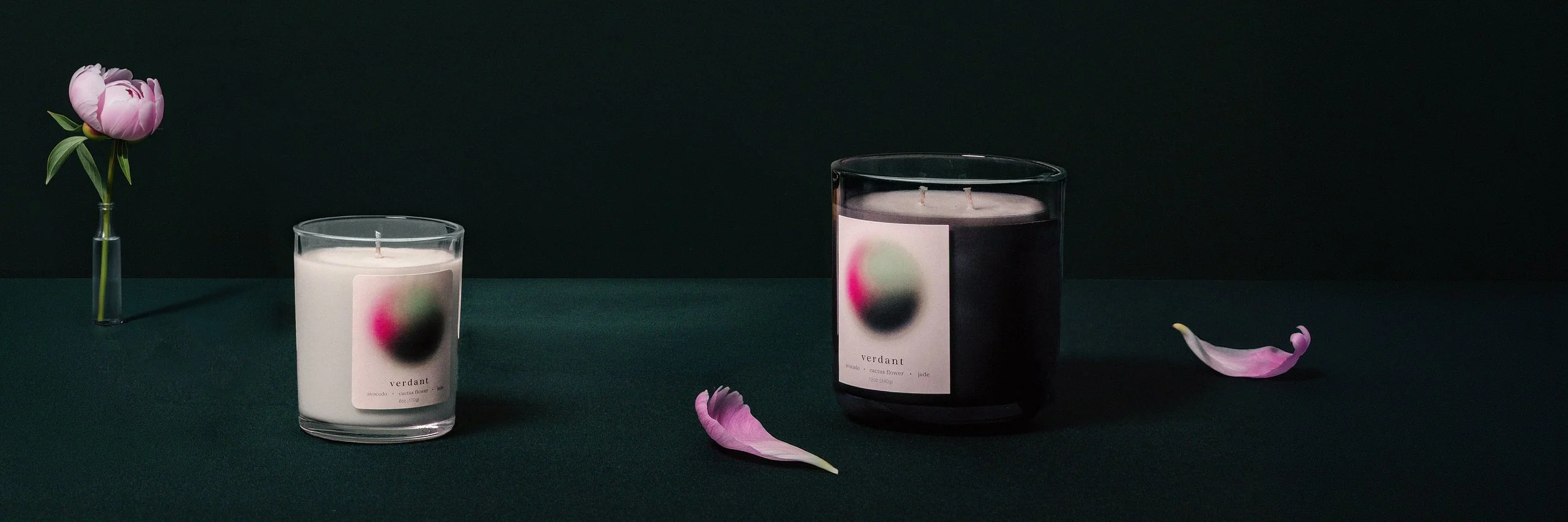 A dark green tabletop with a pink peony in a glass vase on the left, a small white candle in a glass container with a pink and green label in the middle, a larger dark-colored candle with a similar label on the right, and two pink flower petals on the table.