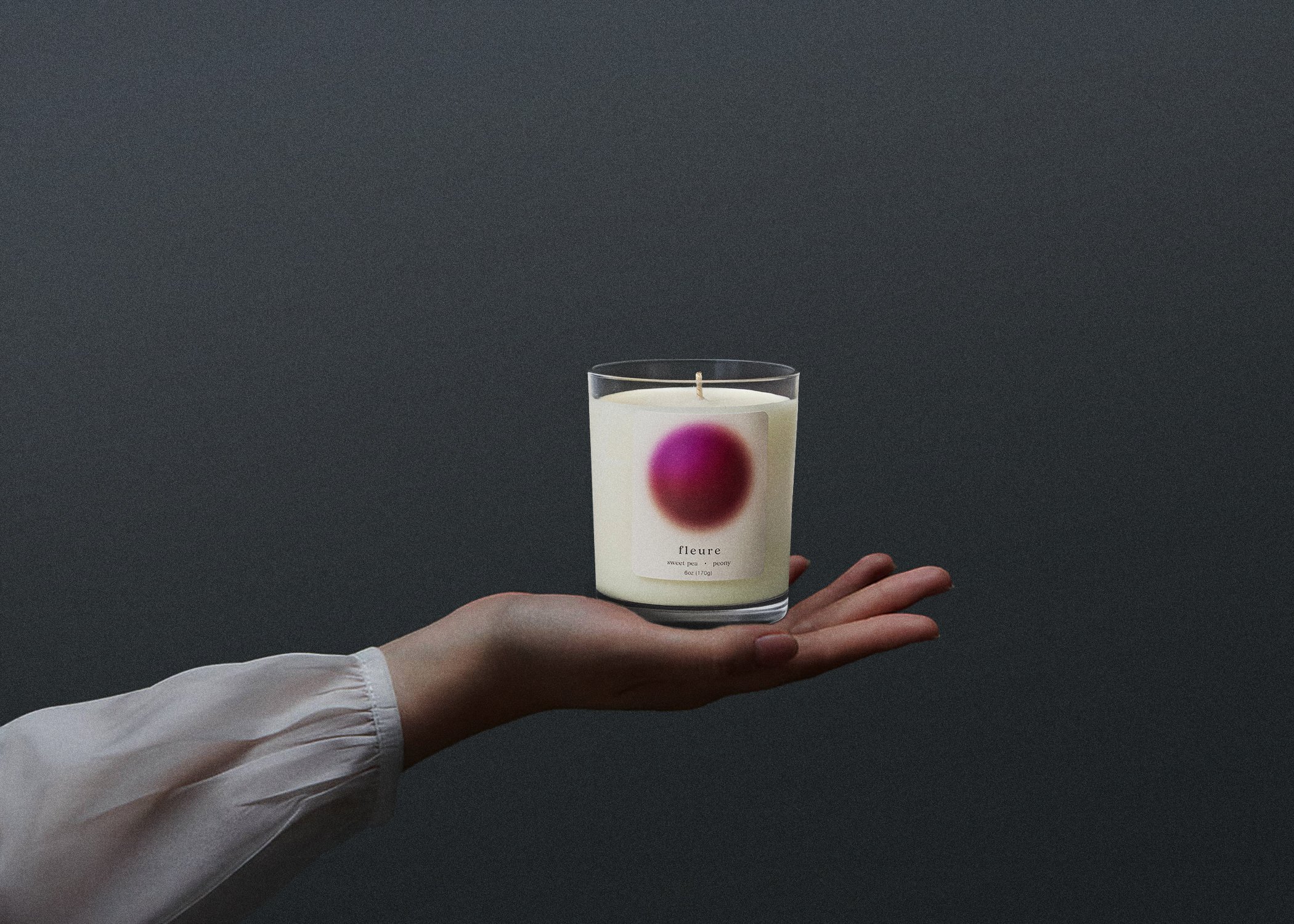 Hand holding a white scented candle in a glass container with a pinkish-red circle design and label reading 'fleure, sweet pea, peony, 6oz (170g).' Dark background.