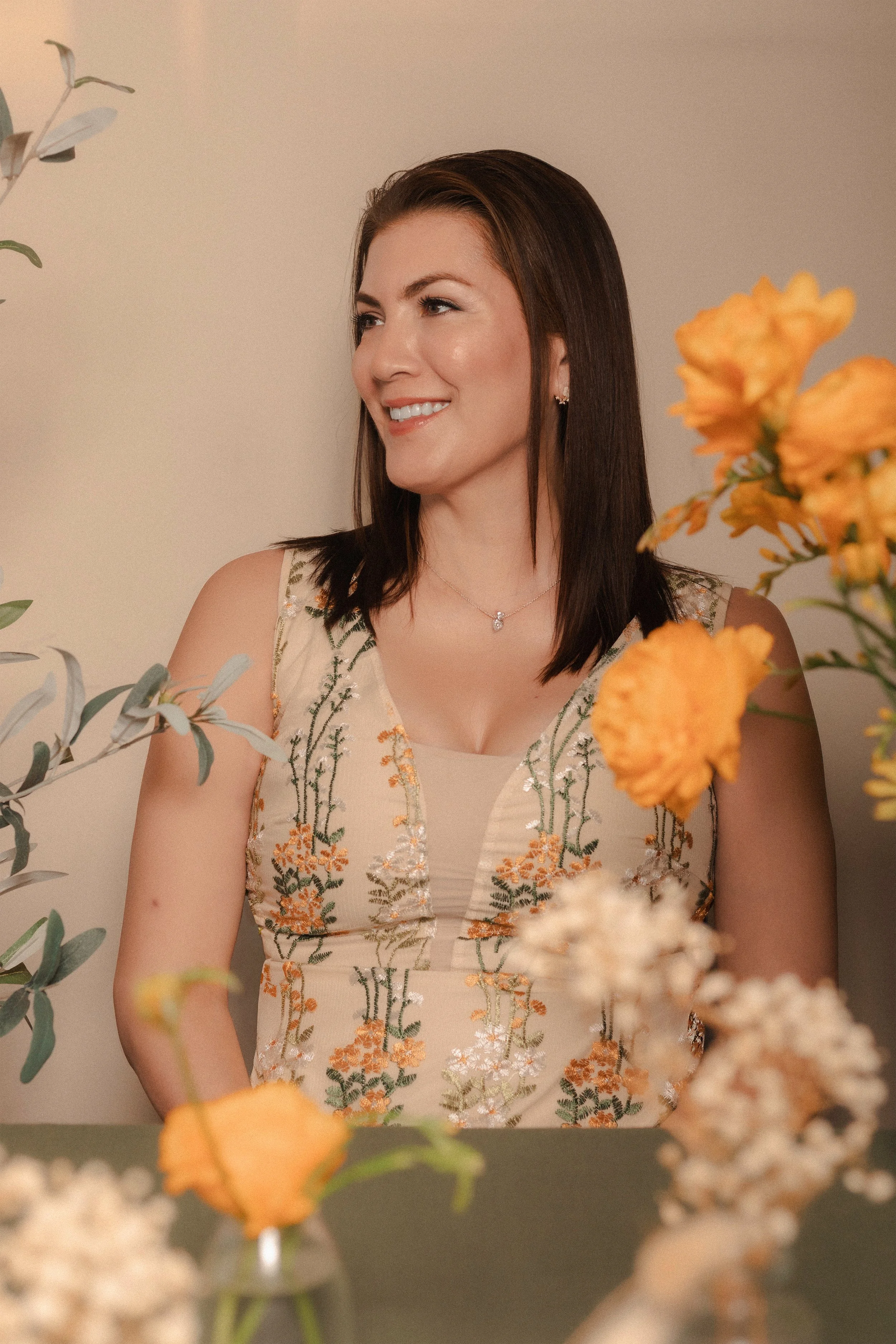 A woman with dark brown hair and a necklace sitting at a table with orange and white flowers, smiling and looking to her left.