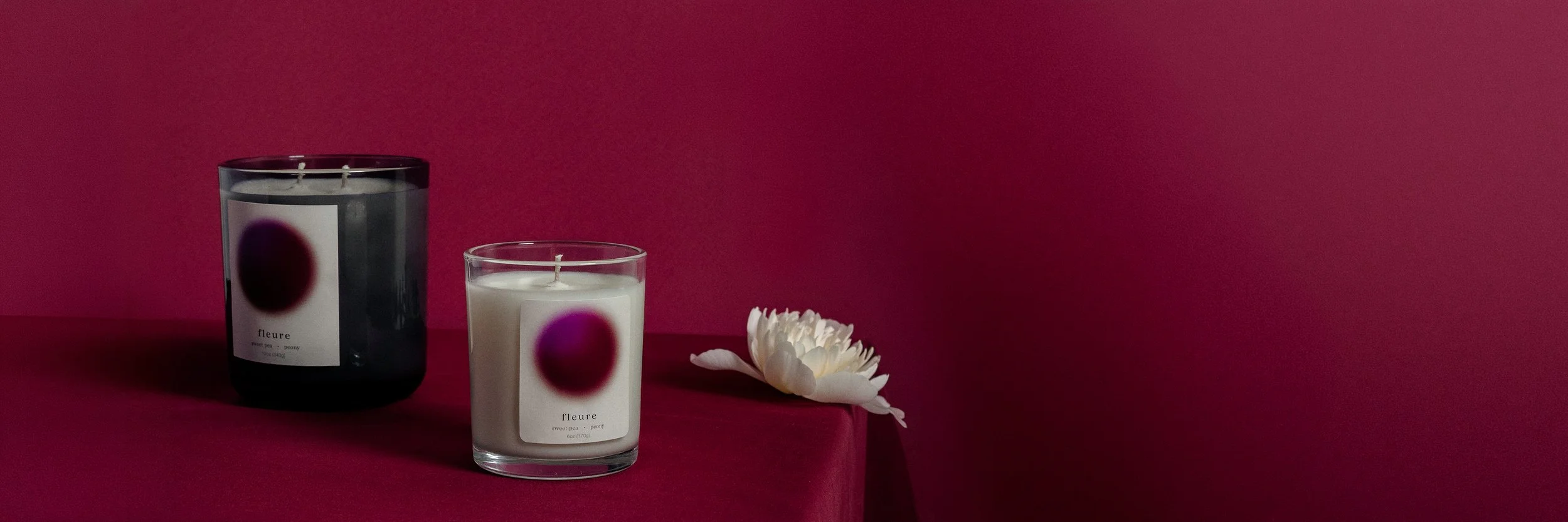 Two fleure scented candles, one in a black jar and one in a clear glass holder, placed on a red surface against a red background, with a white flower beside them.