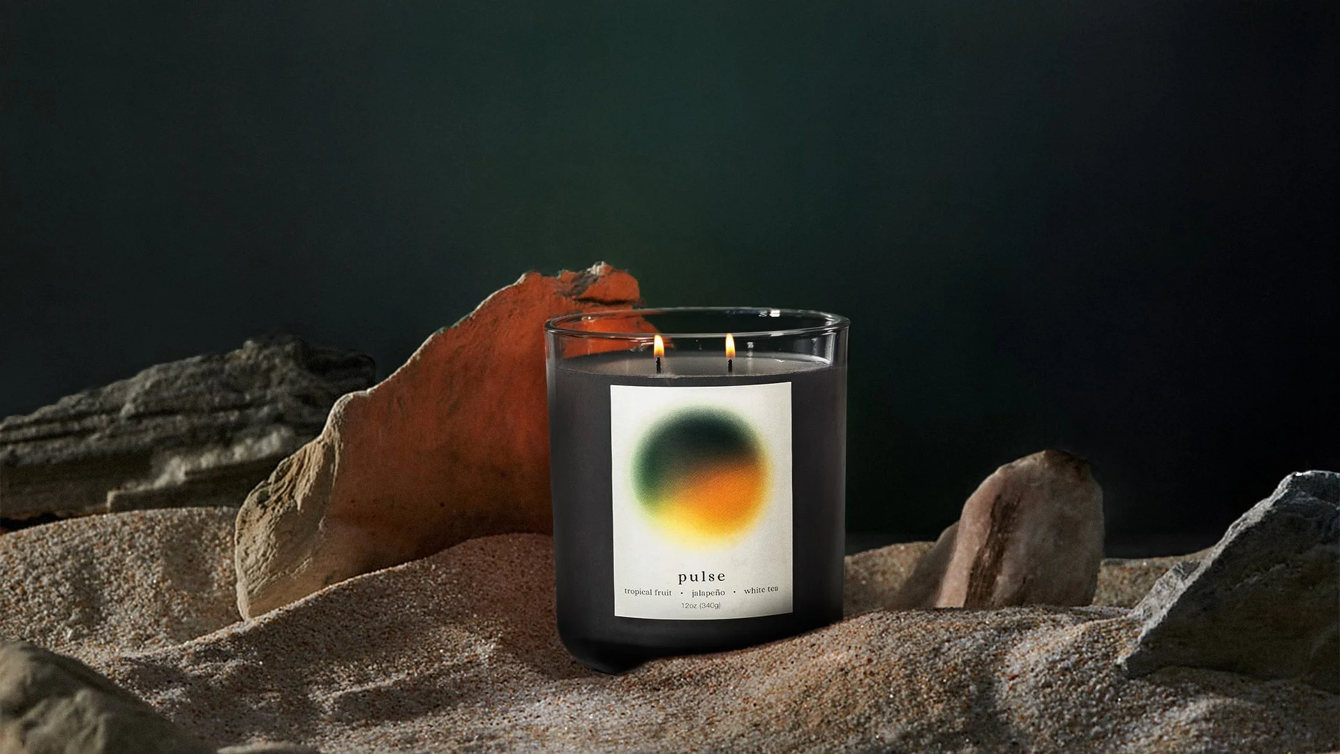 A black candle labeled "pulse" with a colorful abstract design, placed on sand among rocks, with a dark background.