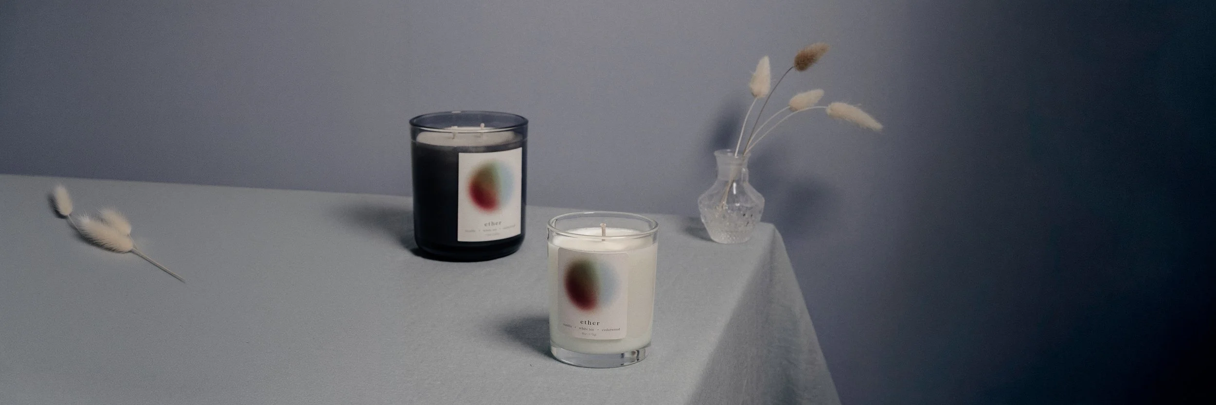 Two candles in glass containers on a white surface with a light blue wall in the background. The candles have abstract art labels, one labeled 'ether'. A small clear vase with dried decorative grasses is placed on the right side of the candles.