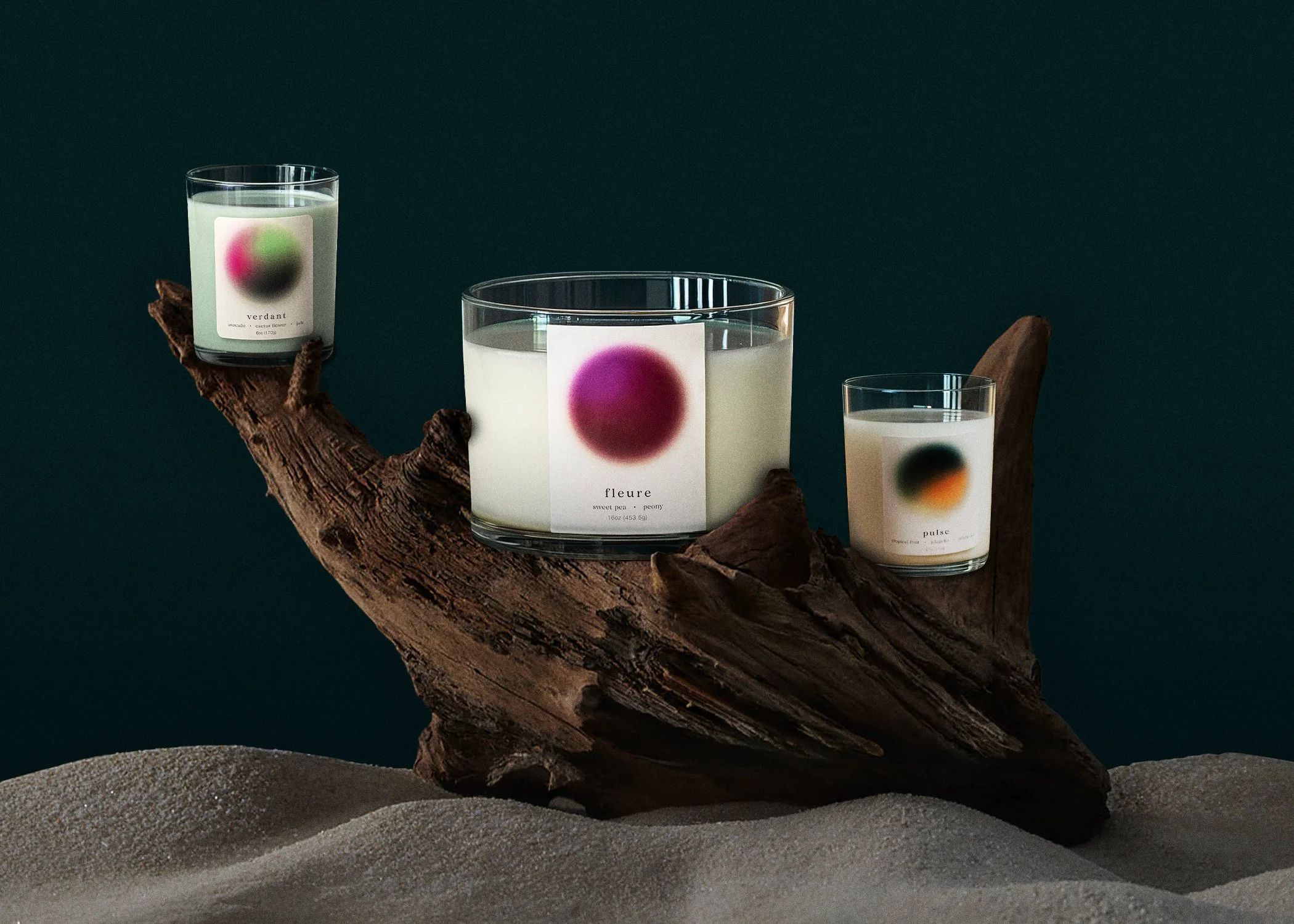 Three candles with modern labels, placed on a piece of driftwood, set against a dark background and sand.