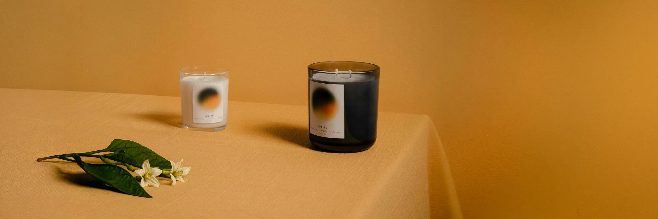 Two candles, one white and one black, with blurred labels, placed on a yellow table against a matching yellow background, with a small cluster of green leaves and white flowers in the foreground.