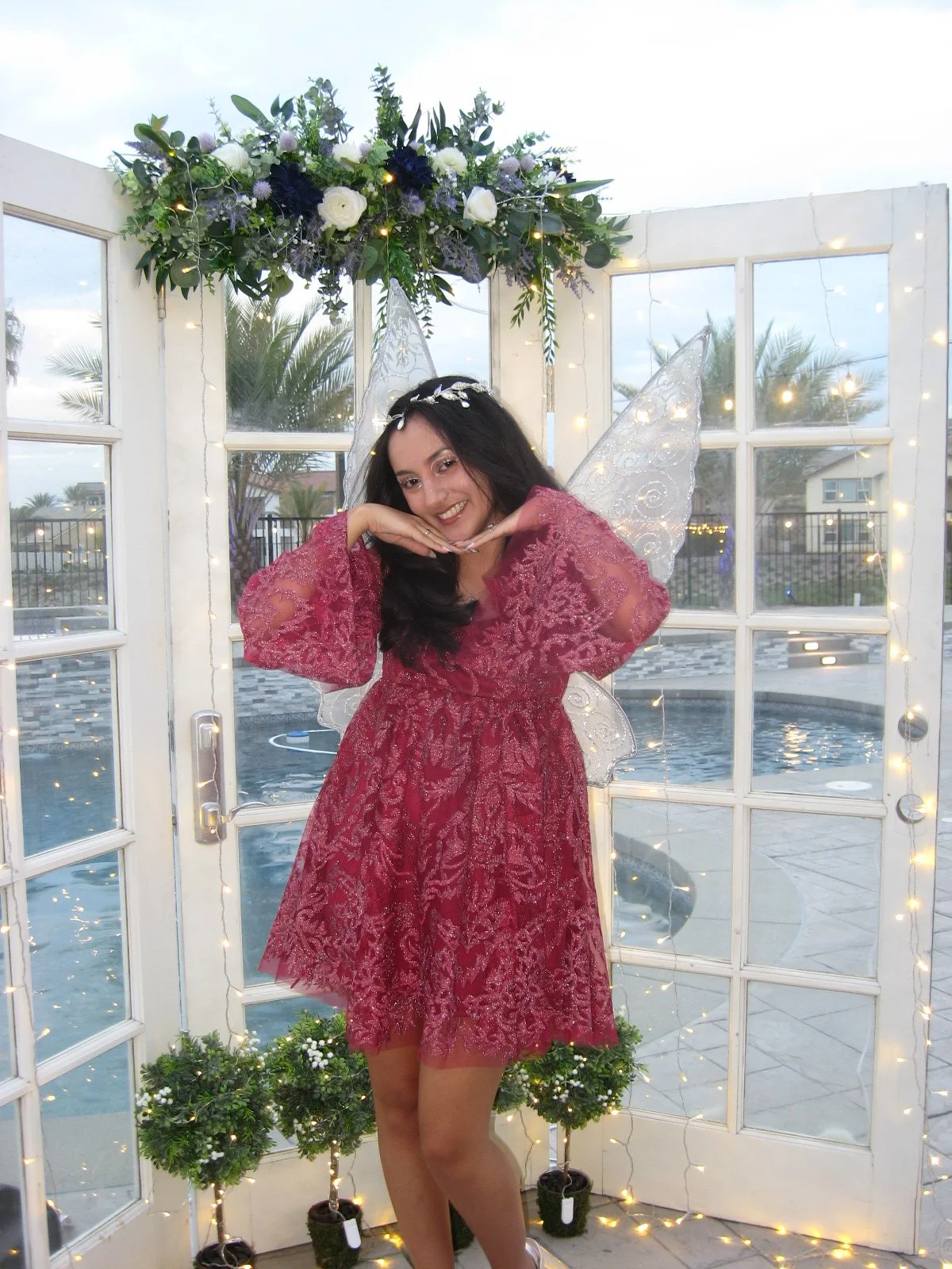 girl in red sparkly dress with fairy wings, smiling with pool and french doors in the background