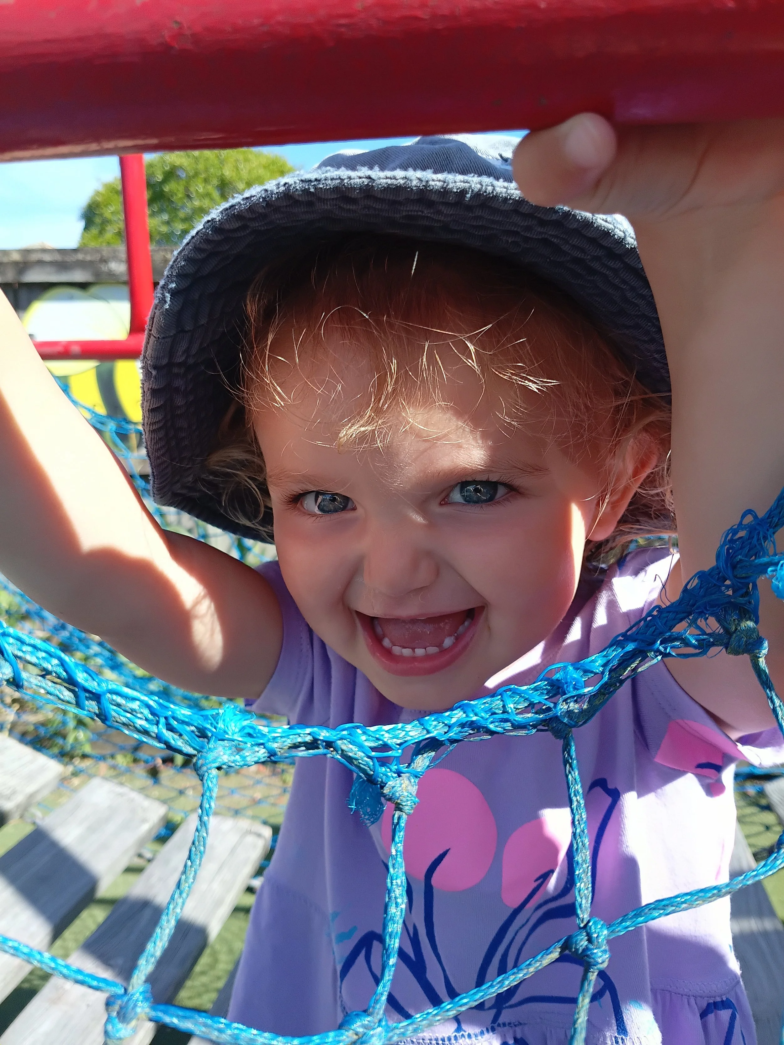 Young girl with blond hair and blue eyes smiling, wearing a dark sunhat and a purple shirt with pink and blue design, playing behind a blue playground net.