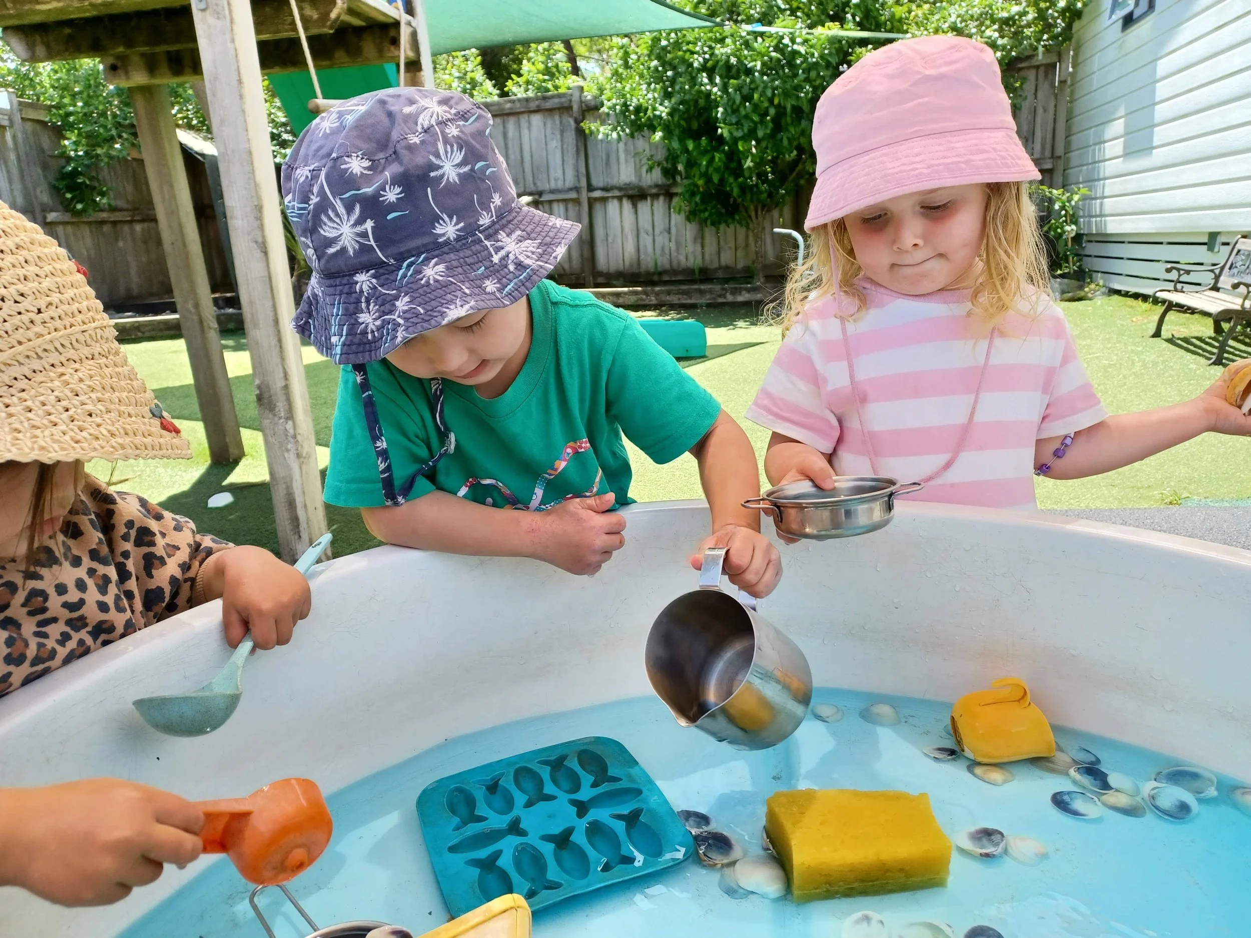 Three young children playing with water toys in a backyard sandbox during daytime. They wear colorful hats and are engaged with cups and scoops in the water.