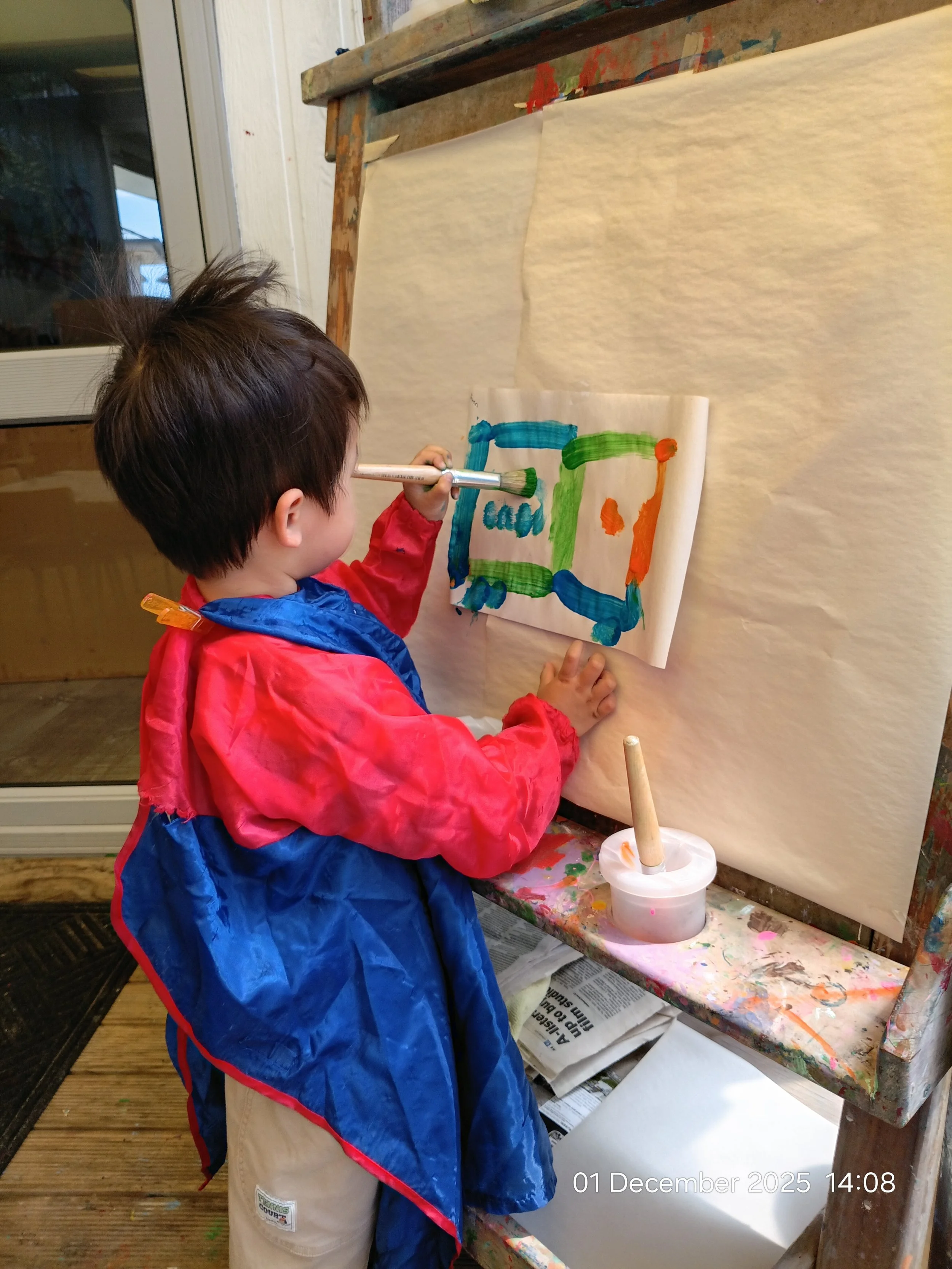 A young child with brown hair painted with a blue and pink smock, standing at an easel and painting on paper with colorful brushes, in an indoor art area.