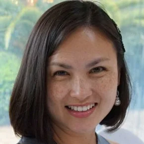 Close-up of a woman with dark hair and earrings smiling outdoors.