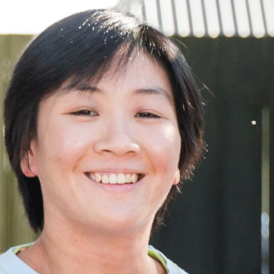 Close-up of a young woman smiling outdoors with a background of a house and a wooden fence.