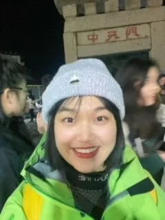 A woman outdoors at night smiling, wearing a gray beanie and a bright green jacket, with other people and a building in the background.