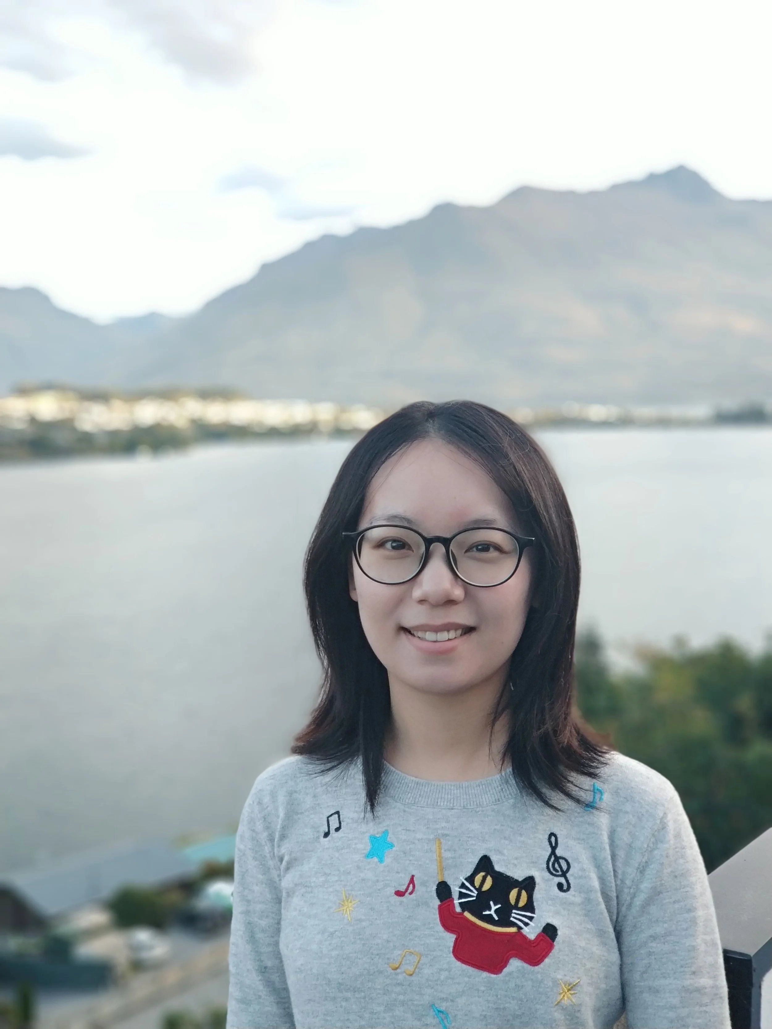 A young woman with shoulder-length dark hair, wearing glasses and a gray sweatshirt with a black cat playing a violin and colorful musical notes, standing outdoors near a body of water with mountains in the background.
