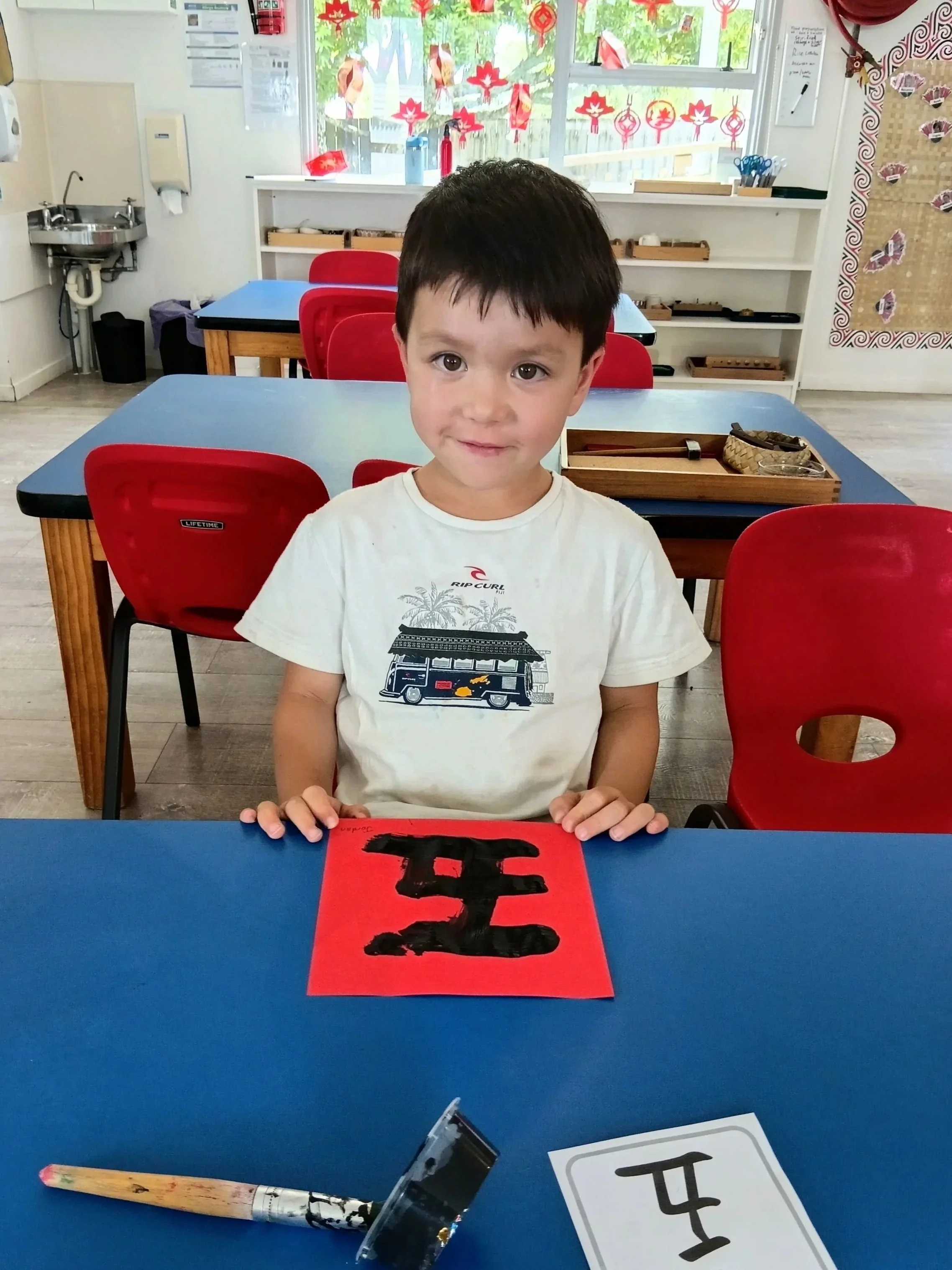 A young boy sitting at a blue table in a classroom, holding a red paper with a black Chinese character painted on it. There is a white card with the same character on the table, along with a paintbrush and a paint container. The classroom has red cha