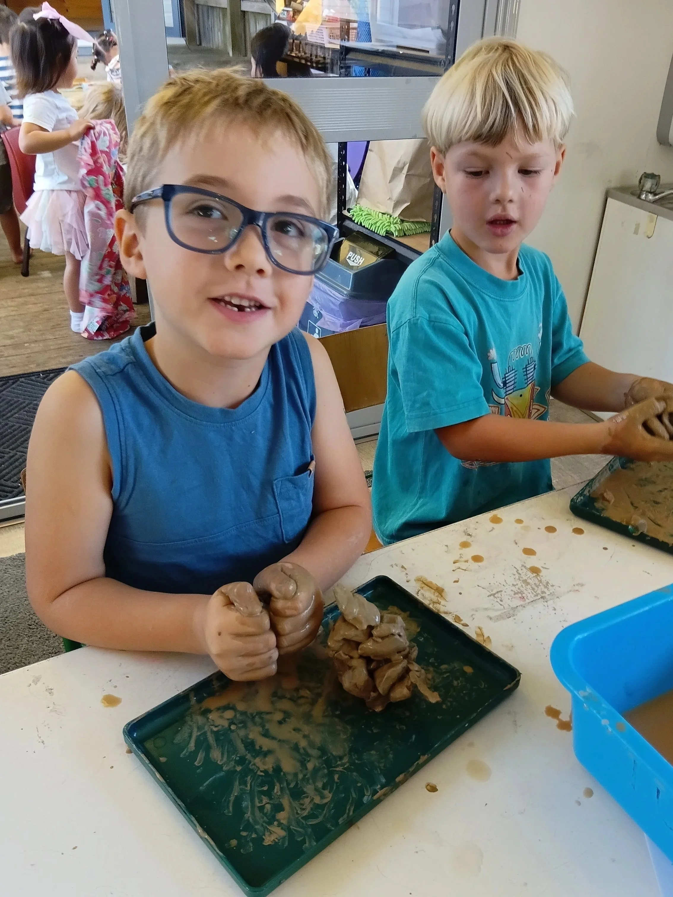 Two young boys are playing with brown dough at a table in a classroom or play area. They appear to be engaged in a sensory activity, with their hands covered in the dough and kneading or shaping it. Other children are visible in the background.