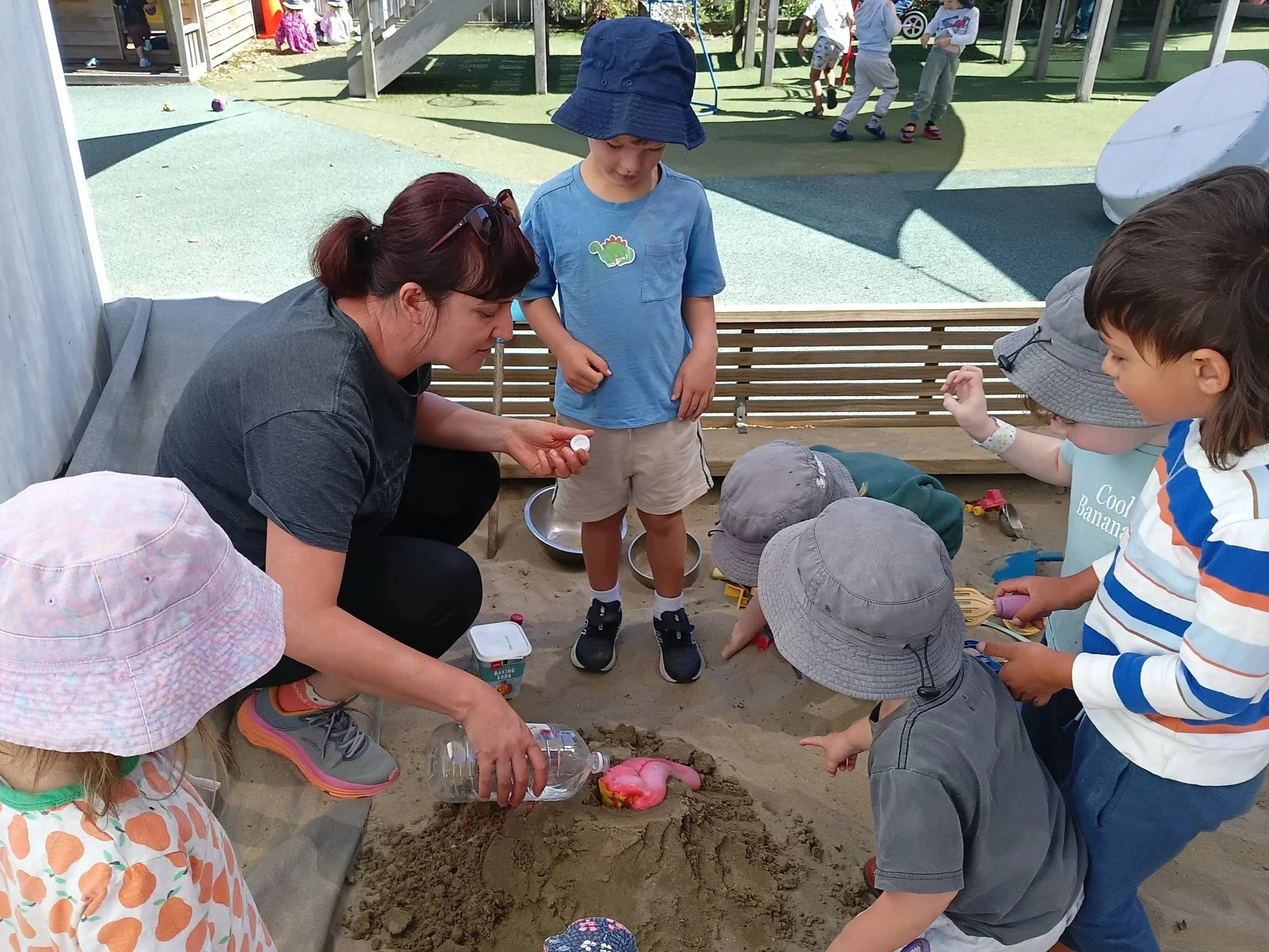 Adults and children at a sandbox playground, some children wearing hats, playing with a pink plastic toy and exploring the sand while an adult pours water into the sandbox.