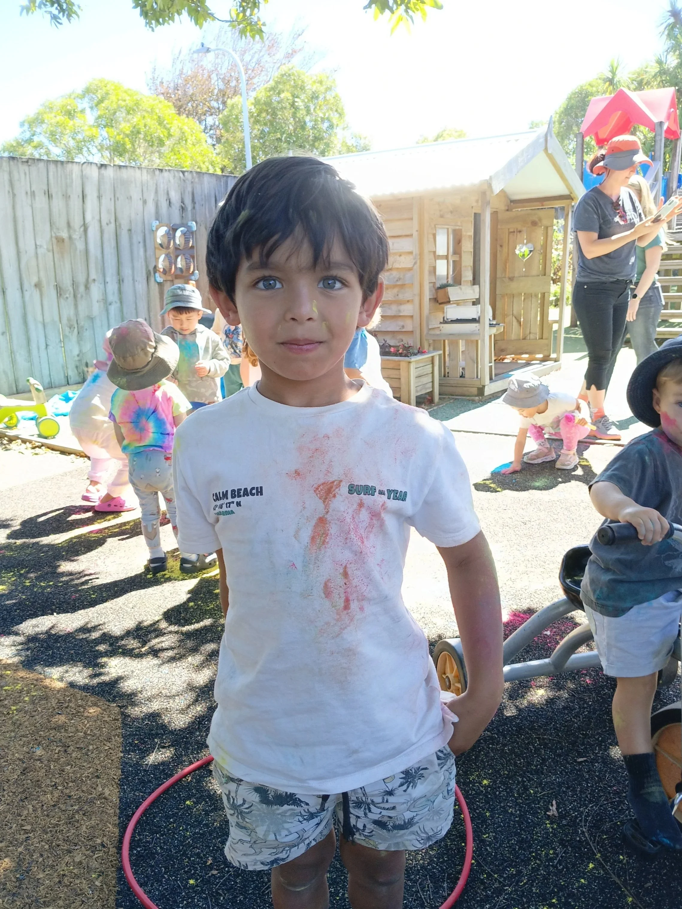 A young boy with dark hair and light skin standing inside a pink hula hoop, outdoors at a playground with other children and adults in the background, some wearing hats.