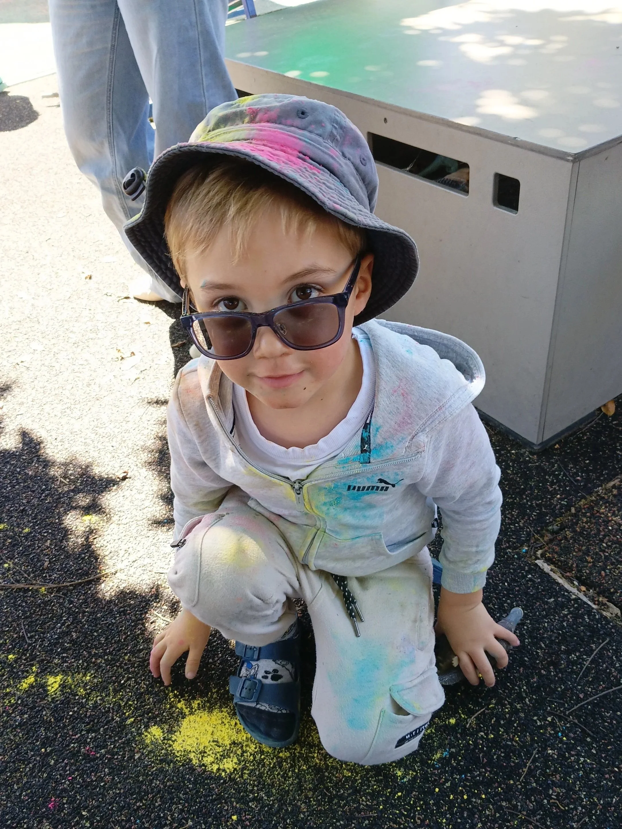 Young boy wearing sunglasses and a bucket hat, squatting on the ground, with colorful powder on his clothes, likely at a color festival or celebration.